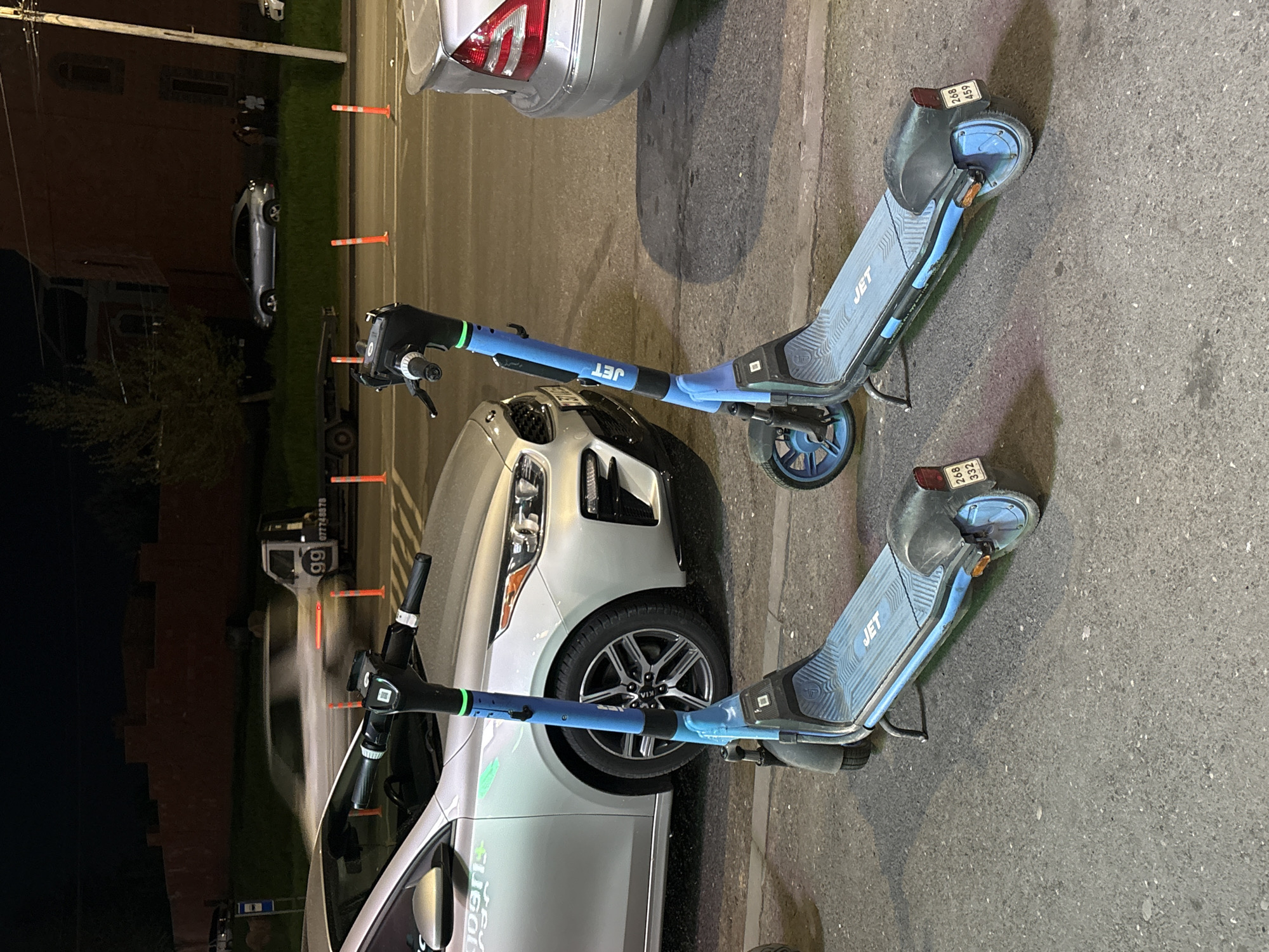 The image captures a nighttime street scene in Yerevan, Armenia, dominated by two blue and black "JET" electric scooters parked on an asphalt surface. They are positioned next to a silver Kia car, which features "sugot" (likely Yugot, a car-sharing service) branding on its side. The Kia's headlights and grille are visible, as well as its wheel with a "KIA" logo on the hubcap.
In the background, a darker silver car is parked on a grassy verge, lined with several orange traffic cones. A large, dark building, possibly residential or commercial, stands behind the grass. Further down the street, a white van is partially visible, displaying "99" and a phone number "077749878".
The scooters have visible identification numbers on their rear fenders: one reads "268 459" and the other "266 332". The brand "JET" is clearly printed on both scooters. The scene is illuminated by artificial light sources, indicating it is dark outside. The ground appears dry. No active human subjects are clearly discernible in the foreground, suggesting a quiet urban setting during the evening hours.