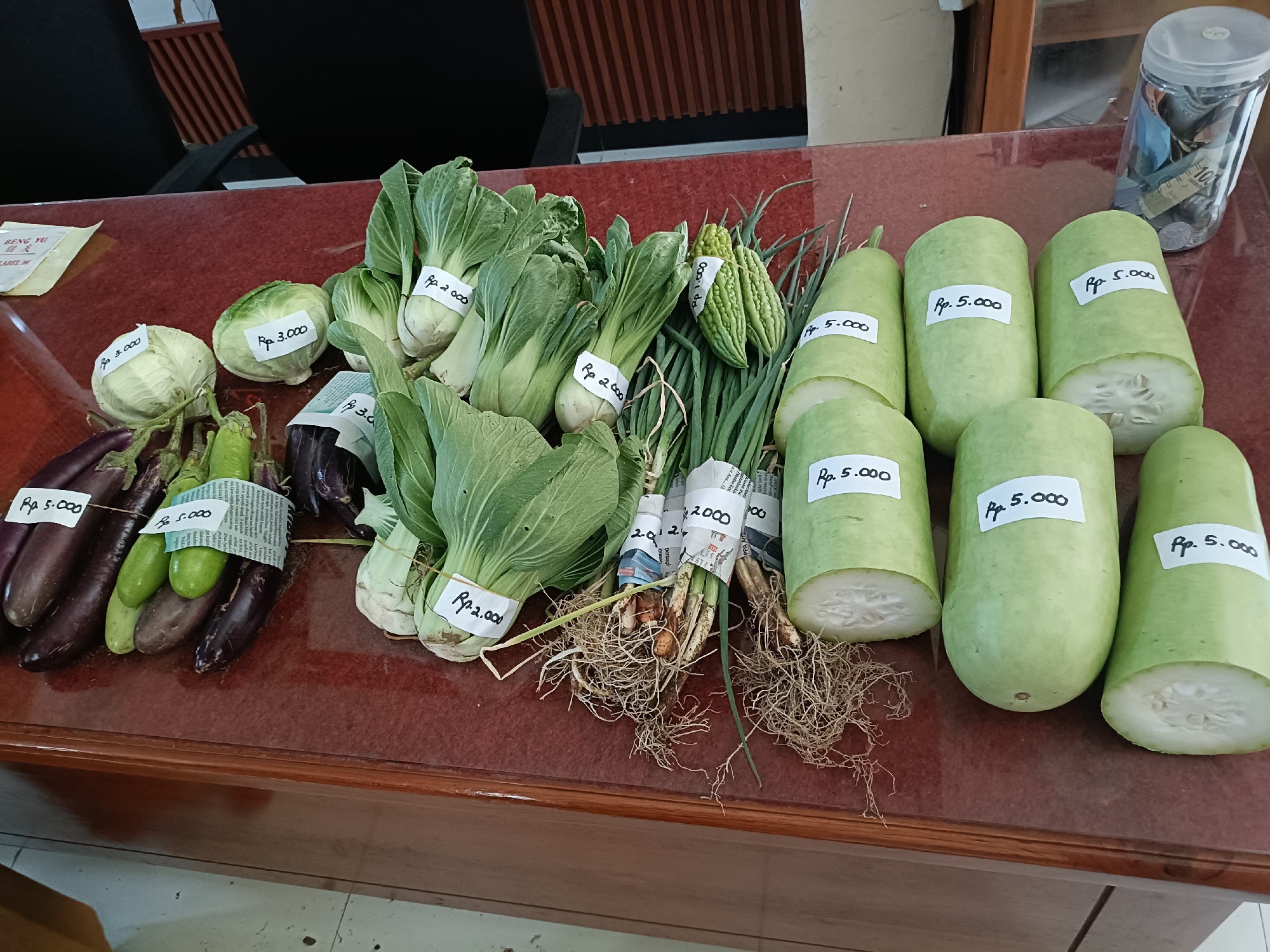 A collection of fresh produce is displayed on a dark reddish-brown table. The items are arranged in groups and have handwritten price tags attached. From left to right, there are several dark purple eggplants and some green oblong vegetables, possibly cucumbers or gourds. Next to these are bundles of leafy green vegetables, likely bok choy or similar greens, with tags indicating "Rp. 2.000" and "Rp. 3.000". There are also bitter gourds with price tags of "Rp. 4.000". Bunches of green onions with visible roots are present, wrapped in newspaper with handwritten prices. Finally, a row of large, light green, cylindrical vegetables, likely gourds or squash, are displayed, each with a tag indicating "Rp. 5.000". In the background, a clear plastic container filled with money and coins is visible on the right side of the table. A hint of a wooden structure is present in the upper background, and a white packet with the text "BENG YU" is visible on the far left. The scene appears to be an outdoor market stall or a produce stand in Kediri City, Indonesia, given the currency denomination and likely context.