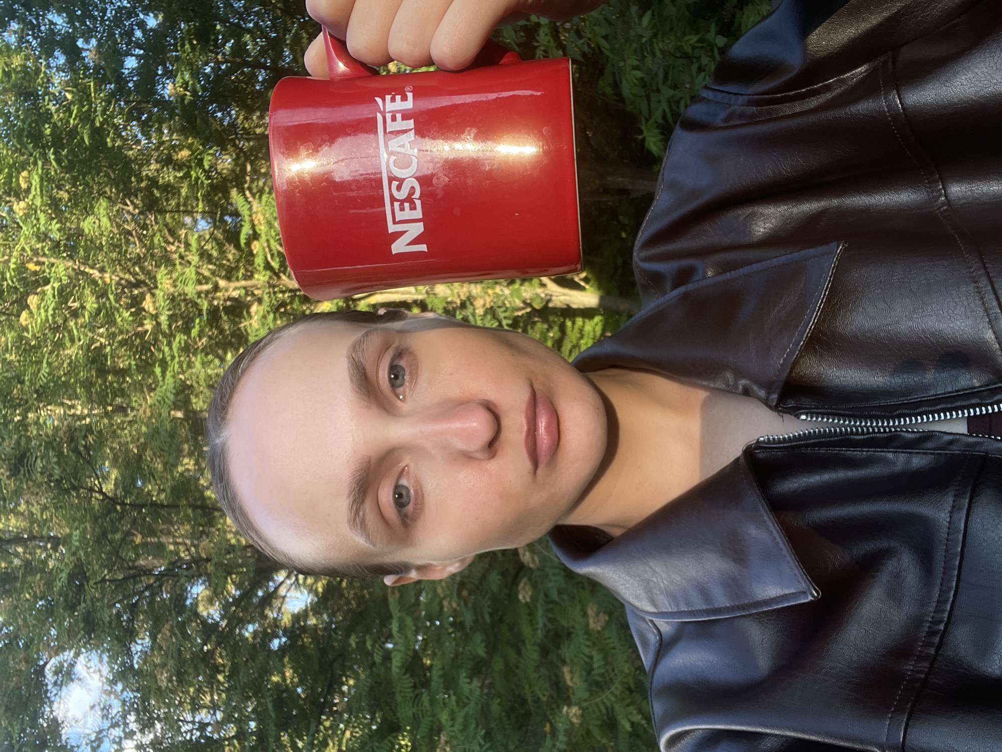 The image, rotated 90 degrees clockwise from its natural orientation, presents a close-up, portrait-style view of an individual outdoors. The person, seen from the chest up, has light-colored eyes and a neutral expression, looking directly at the camera. They are wearing a dark brown or black leather-like jacket with a prominent collar and a silver-toned zipper detail.
The individual holds a bright red mug with a handle, positioned towards the upper right side of the frame. The brand "NESCAFÉ®" is clearly visible in white capital letters printed vertically on the side of the mug, which reflects a strong gleam of sunlight.
The background is filled with lush green foliage, likely trees or dense bushes, indicating an outdoor setting in Sharkan, Russia. Patches of blue sky are visible through the leaves. The bright natural light suggests it is daytime, casting both highlights and shadows on the subject and objects. No specific activity beyond holding the mug is discernible, but the scene conveys a relaxed outdoor moment.