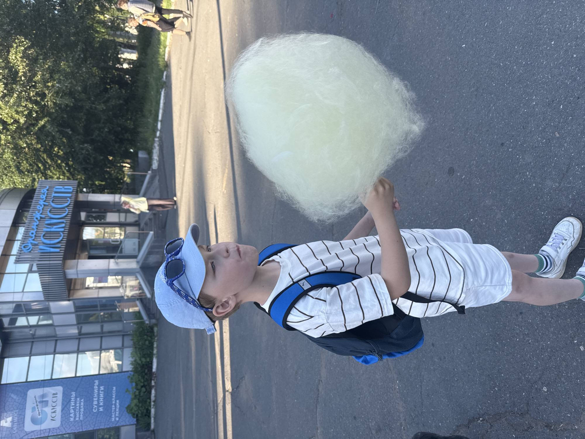 The image displays an outdoor scene in Izhevsk, Russia, featuring a young boy consuming cotton candy with the "Оранжерея Искусств" (Orangery of Arts) building in the background.
**Location:** The building identified as "Оранжерея Искусств" (Orangery of Arts) is visible in the background, located in Izhevsk, Russia. A banner attached to the building lists services including "Картины, Выставки, Продажа" (Pictures, Exhibitions, Sale), "Сувениры и Книги" (Souvenirs and Books), and "Мастер-классы и Лекции" (Master Classes and Lectures).
**Main Subject:** A boy, estimated to be between 6 and 9 years old, stands in the foreground, facing slightly to his right. He wears a light blue baseball cap with blue-framed sunglasses perched on the brim, a white t-shirt with thin horizontal brown stripes, white shorts, light green and white striped socks, and white sneakers with Velcro fasteners. A dark blue backpack with light blue accents is worn on his back. The boy holds a large, pale yellow-green mass of cotton candy with both hands, raised to his mouth, and appears to be in the process of eating it, with his cheeks slightly puffed.
**Background People:**
* To the left, near the building's entrance, a woman wearing a light-colored top and dark pants is standing.
* To the far right, two individuals are walking away from the viewer on a paved surface.
**Environment:** The ground is dark asphalt pavement. The scene is illuminated by natural daylight, with shadows cast to the left of the boy, indicating a light source from the right. The "Оранжерея Искусств" building is a modern structure with a glass facade. Trees and green foliage are visible beyond the building and to its right.