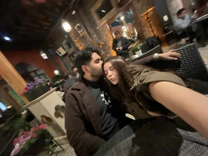 A man kisses a woman's head in an outdoor setting. The man is wearing a brown coat over a black turtleneck. The woman reclines on a wicker chair, her eyes closed. Her arm is extended in front. Other patrons are visible in the background. A flower box is situated nearby. The photo has a slightly tilted angle.