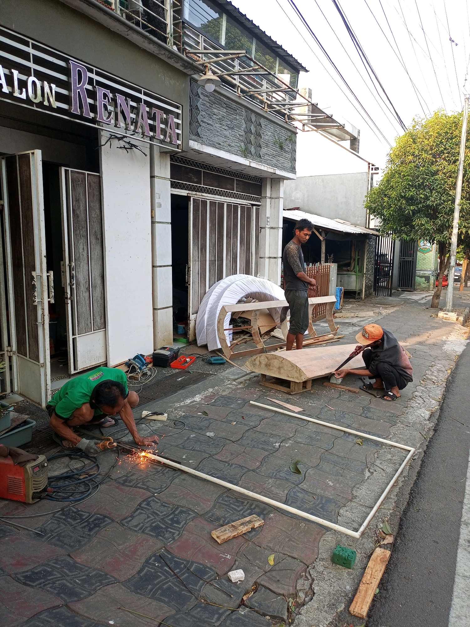 The image depicts an outdoor scene in Batu, Indonesia, where three individuals are engaged in construction work. The setting appears to be a sidewalk in front of a business labeled "SALON RENATA."
In the foreground, a man wearing a green t-shirt and dark shorts is crouching and welding a white pipe, with sparks flying from the welding point. He is wearing gloves. To his right, another man, wearing a dark hooded jacket and dark pants, is squatting and sawing a curved wooden board. He has an orange hat on. A third man, wearing a dark t-shirt and dark pants, is standing and observing the work.
Various construction materials and tools are scattered around the men. This includes the wooden boards, the white pipes, a welding machine, an electric saw, and other assorted items. In the background, there is a partially constructed white dome-like structure, possibly a shelter or a decorative piece. The building behind them has a stone facade and metal window frames. Beyond the building, trees and power lines are visible, indicating an urban or semi-urban environment. The ground is paved with interlocking blocks. The overall impression is one of active construction or craftwork taking place on a public walkway.