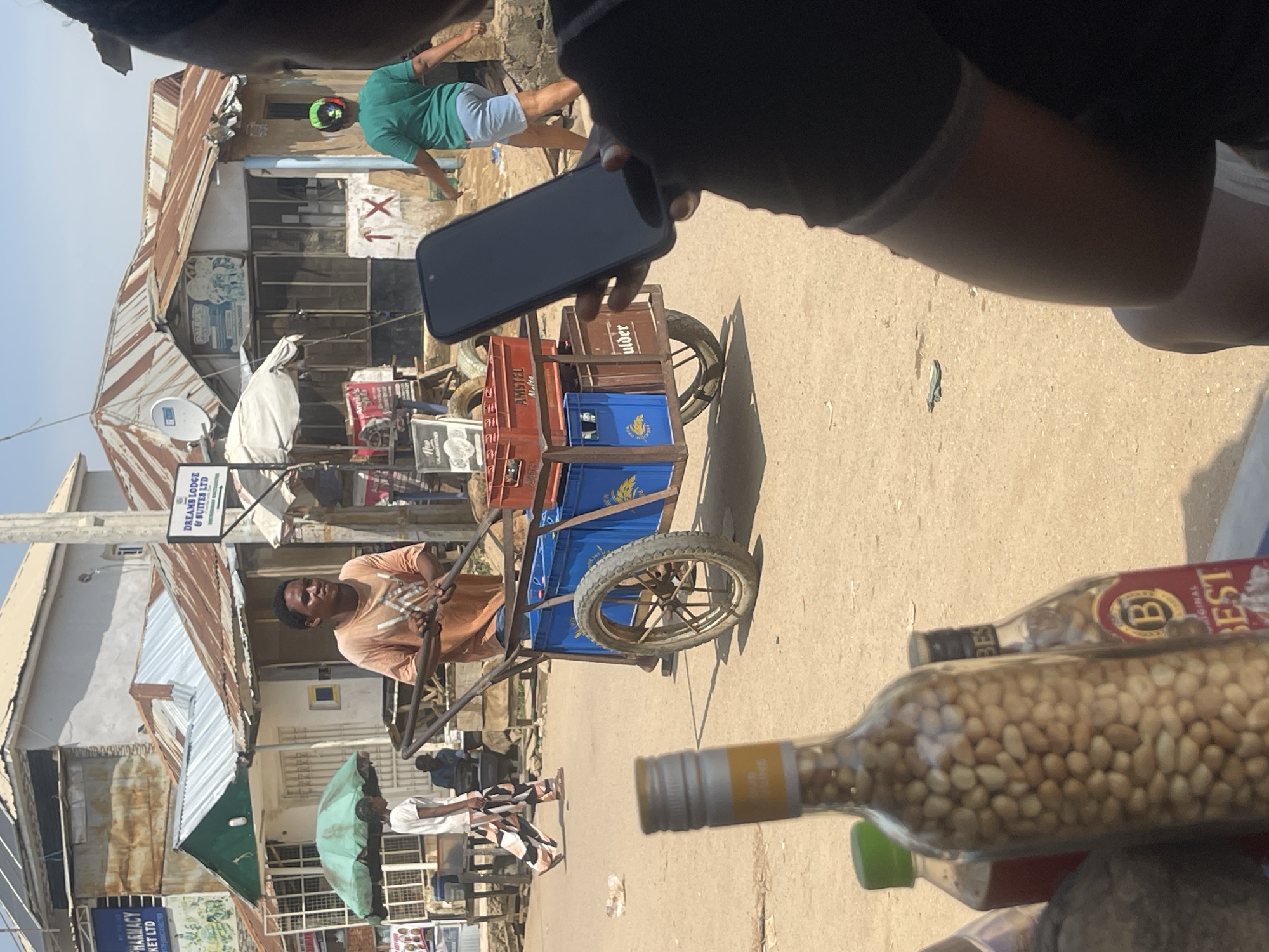 The image depicts a street scene in Abuja, Nigeria. In the foreground, a person is holding a dark smartphone, obscuring a portion of their face and shoulder. Behind them, a man in an orange t-shirt pushes a two-wheeled cart laden with crates of beverages. The cart has blue plastic crates and some brown crates, one of which is labeled "Amstel Malta" and another partially visible label reads "uider". To the left, a woman in a white t-shirt and patterned pants walks away from the camera, and another person in a green t-shirt and blue shorts walks towards the right side of the frame. Several buildings with corrugated metal roofs line the street, with signs for "Dreams Lodge & Suites Ltd." and "Palmas" visible. The ground is sandy and dusty. In the immediate foreground, several bottles are visible, including one filled with peanuts, and another with a red liquid. The overall atmosphere suggests a bustling market or commercial area.