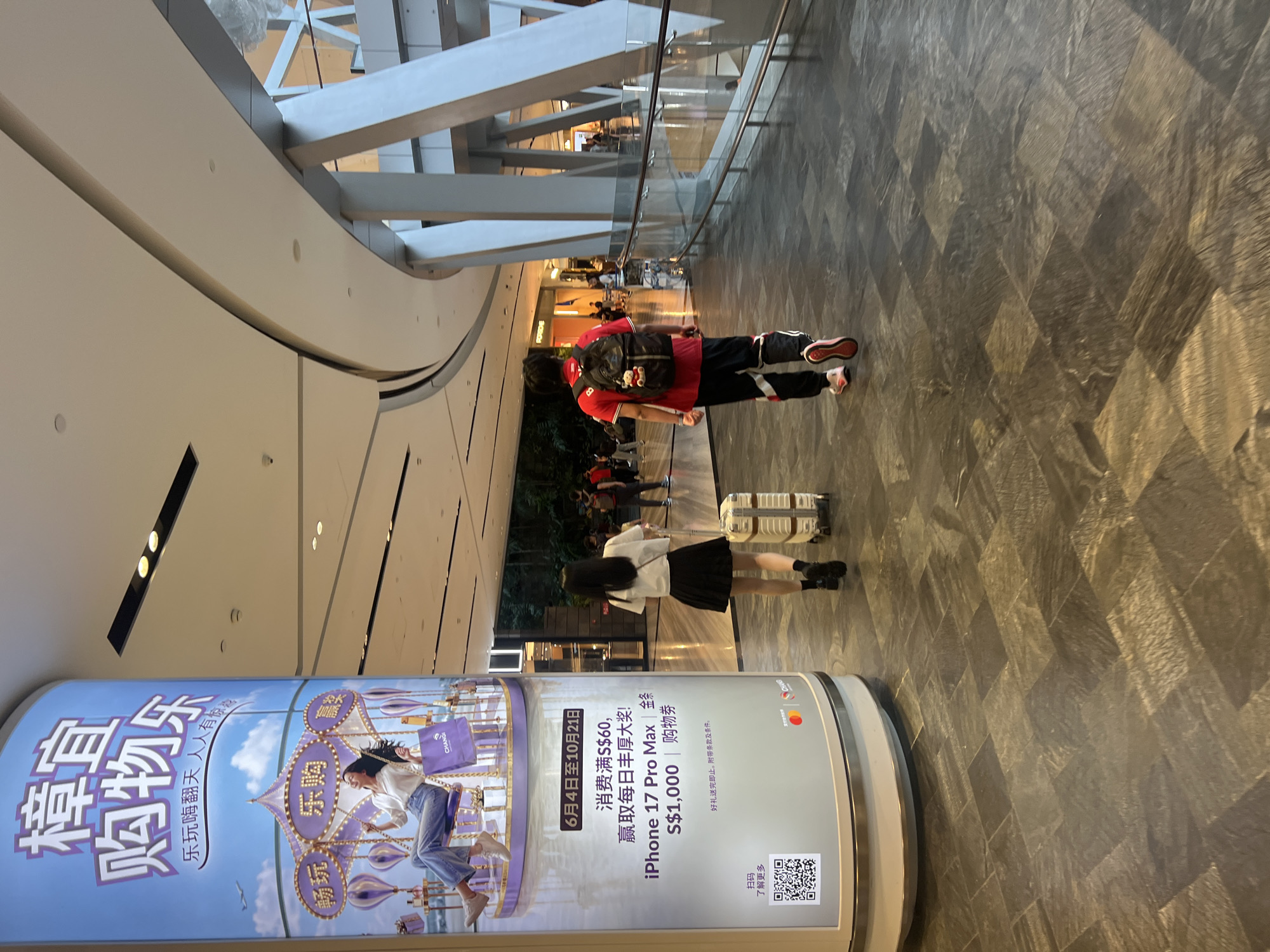In Singapore, at the Changi Airport Terminal 3 Departure Hall, a young woman with long black hair, wearing a white t-shirt and a black skirt, is seen from behind, pulling a white suitcase with a small brown teddy bear keychain hanging from its handle. She is walking away from a large cylindrical advertising pillar. The pillar features an advertisement in Chinese, promoting a shopping event with a carousel graphic and text including "iPhone 17 Pro Max" and a "S$1,000" prize. To her right, a young man with short dark hair, wearing a red t-shirt with black accents, black pants with white stripes, and a black backpack, is also walking away from the pillar, presumably in the same direction. He has a small keychain hanging from his backpack as well. They are both walking on a dark grey tiled floor. In the background, other travelers are visible, some walking and others standing. The environment is brightly lit, with modern architectural elements, including curved ceilings and large support columns.