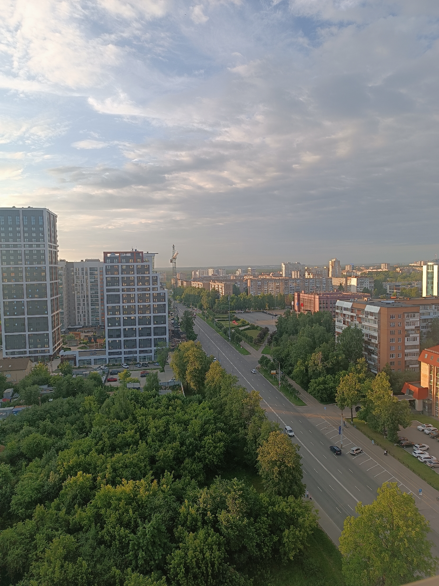 This is a high-angle, wide shot of a city street scene in Idrisovo, Russia, likely taken during the late afternoon or early evening. The sky is overcast with scattered clouds, allowing some sunlight to filter through, creating soft lighting. In the foreground, a dense canopy of green trees occupies the lower left and right portions of the image, with some lighter, yellow-green foliage visible, suggesting the season is late spring or summer.
A multi-lane asphalt road runs diagonally through the center of the frame, from the upper left to the lower right. Several cars are present on the road, including a white van in the left lane, a dark sedan in the right lane, and a silver sedan further down. A few pedestrians are visible on the sidewalk along the right side of the road. Parking areas with multiple cars are located alongside the road.
On the left side of the road, several modern, high-rise apartment buildings are prominent. The building closest to the foreground is a tall, rectangular structure with a dark facade and numerous windows. Adjacent to it are other apartment buildings of varying heights and architectural styles. A construction crane is visible in the middle ground, behind the buildings.
Further into the background, the cityscape extends with rows of residential buildings, predominantly brick-colored and lighter-toned structures, interspersed with more trees. A few taller, more modern buildings are also visible in the distant skyline. The overall impression is of a developed urban area with a significant amount of green space.