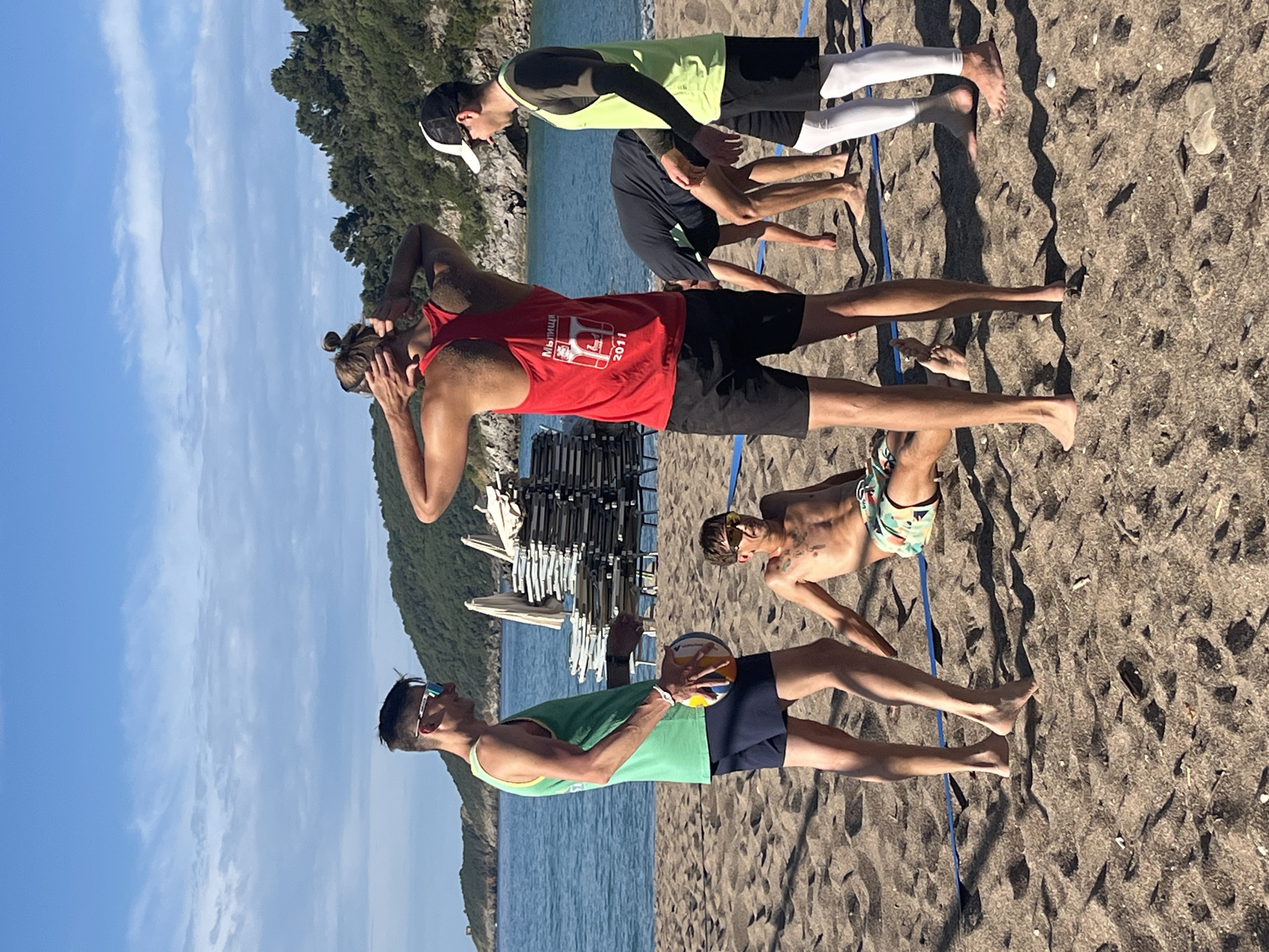 The image depicts a group of five people on a beach. They are standing on sand, with the Adriatic Sea visible in the background, indicating the location is likely a beach in Bečići, Montenegro. A blue volleyball net is stretched across the sand, delineating a playing area.
The people are dressed in casual beach attire including tank tops, shorts, and swim trunks. One person in the foreground is holding a volleyball. Some of the individuals are wearing sunglasses. One is wearing a hat and another appears to be stretching or massaging his head.
Behind the people, there are stacks of beach chairs and sun umbrellas, and beyond that, a rocky shoreline and trees. The sky is clear and blue.