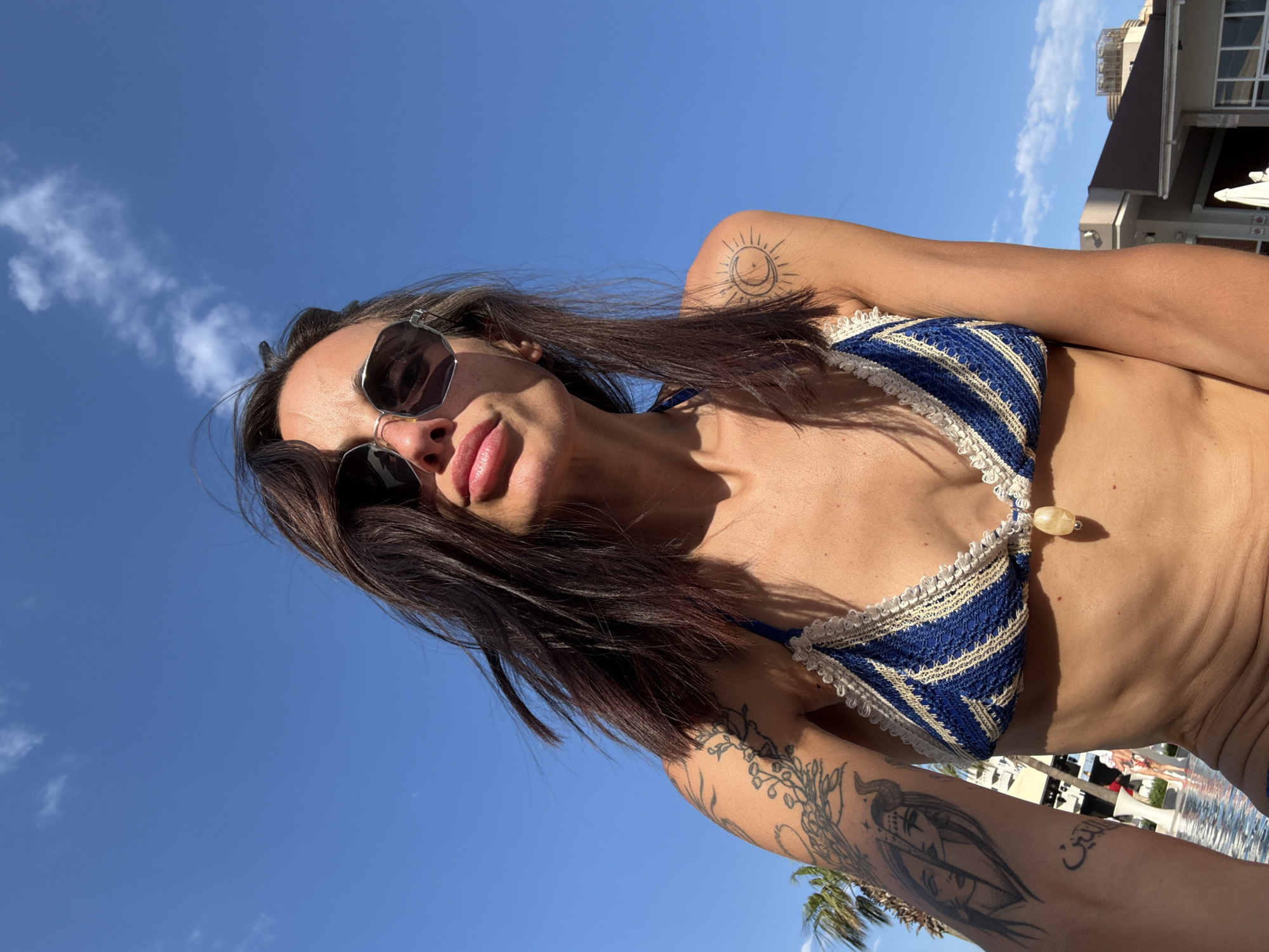 A low-angle, medium shot captures a woman from the chest up, posing for a selfie against a bright blue sky with scattered white clouds. She is wearing a blue and white striped bikini top with a cream-colored crocheted trim. A small, light-colored bead hangs from the center of the bikini top. The woman has dark brown hair that is windswept and partially covers her face. She is wearing large, stylish sunglasses. She has several tattoos on her left arm, including what appears to be a portrait and some floral designs. A tattoo of a sun and crescent moon is visible on her right shoulder. In the background, out of focus, are palm trees, the corner of a building with a brown roof, and a swimming pool. The overall scene suggests a sunny day at a resort or hotel in Aksu, Turkey.