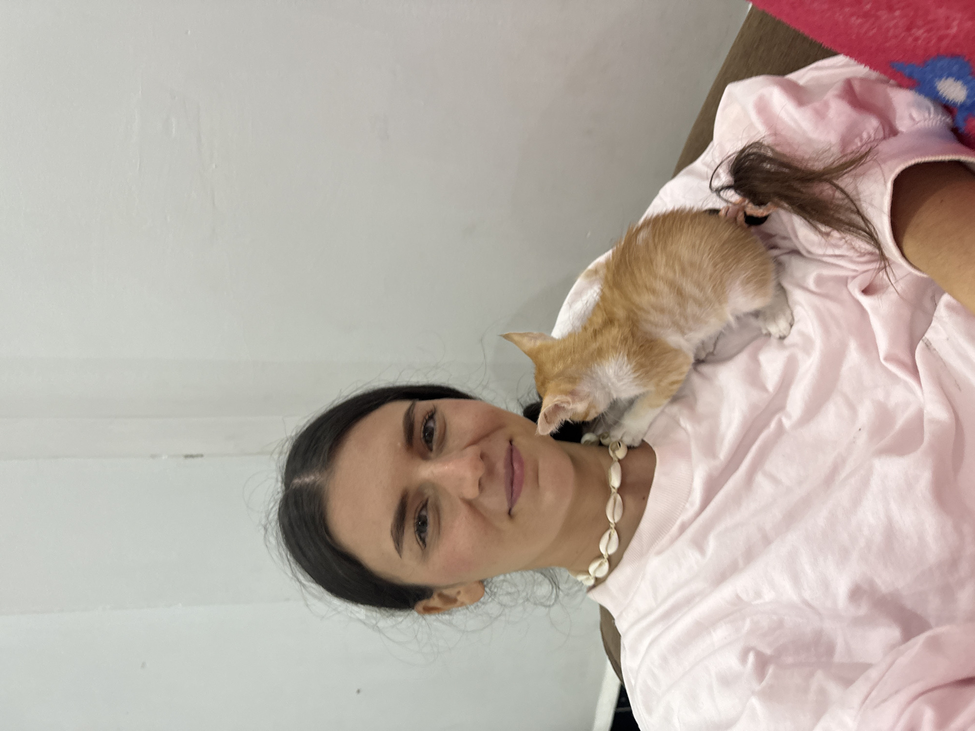 A portrait-oriented image captures a warm interaction between a woman and a small kitten in an indoor setting. The woman, with dark hair, is smiling gently and looking towards the camera. She is wearing a light pink t-shirt and a distinctive cowrie shell necklace. A small ginger and white kitten is comfortably perched on her right shoulder and chest, its head turned downwards near her collarbone.
The background features a plain, light-colored wall, suggesting an indoor environment such as a home. On the right side of the frame, portions of a pink fabric, possibly a blanket or clothing, and a red and blue patterned textile are visible, indicating the woman might be lounging on a bed or sofa. The overall scene conveys a sense of affection and tenderness between the woman and her feline companion. This moment takes place in Mũi Né, Vietnam. There is no visible text within the image.