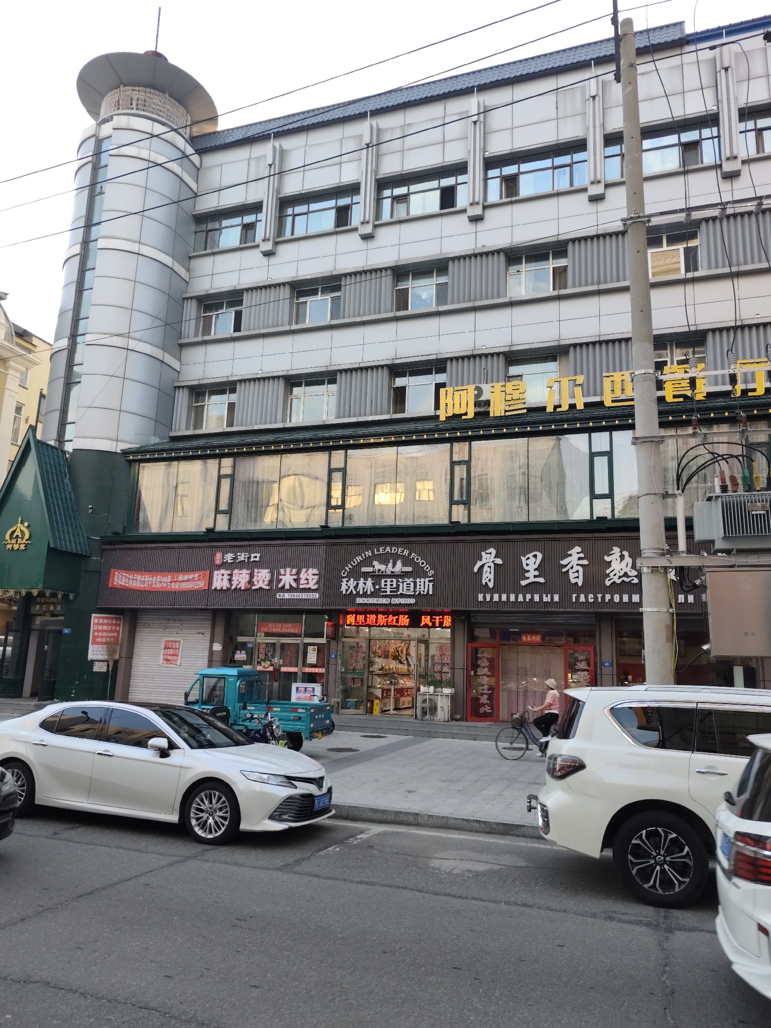 The image shows a street scene in Aihui, China, dominated by a multi-story building with shops on the ground floor. The building's facade is modern, featuring large windows and metallic paneling. A prominent sign in yellow characters is visible on the upper section of the building. Below, the storefronts display various signs, some in Chinese and Russian. One sign reads "CHURIN LEADER FOODS" and another, in Cyrillic, appears to be "КУЛИНАРНЫИ ГАСТРОНОМ" (Culinary Gastronome). In front of the shops, several vehicles are parked. A white sedan is on the left side of the road, and a white SUV is partially visible on the right. A small turquoise truck is parked near one of the storefronts. A person on a bicycle is seen riding on the sidewalk in front of the shops. The environment suggests a commercial district with a mix of businesses and street life.