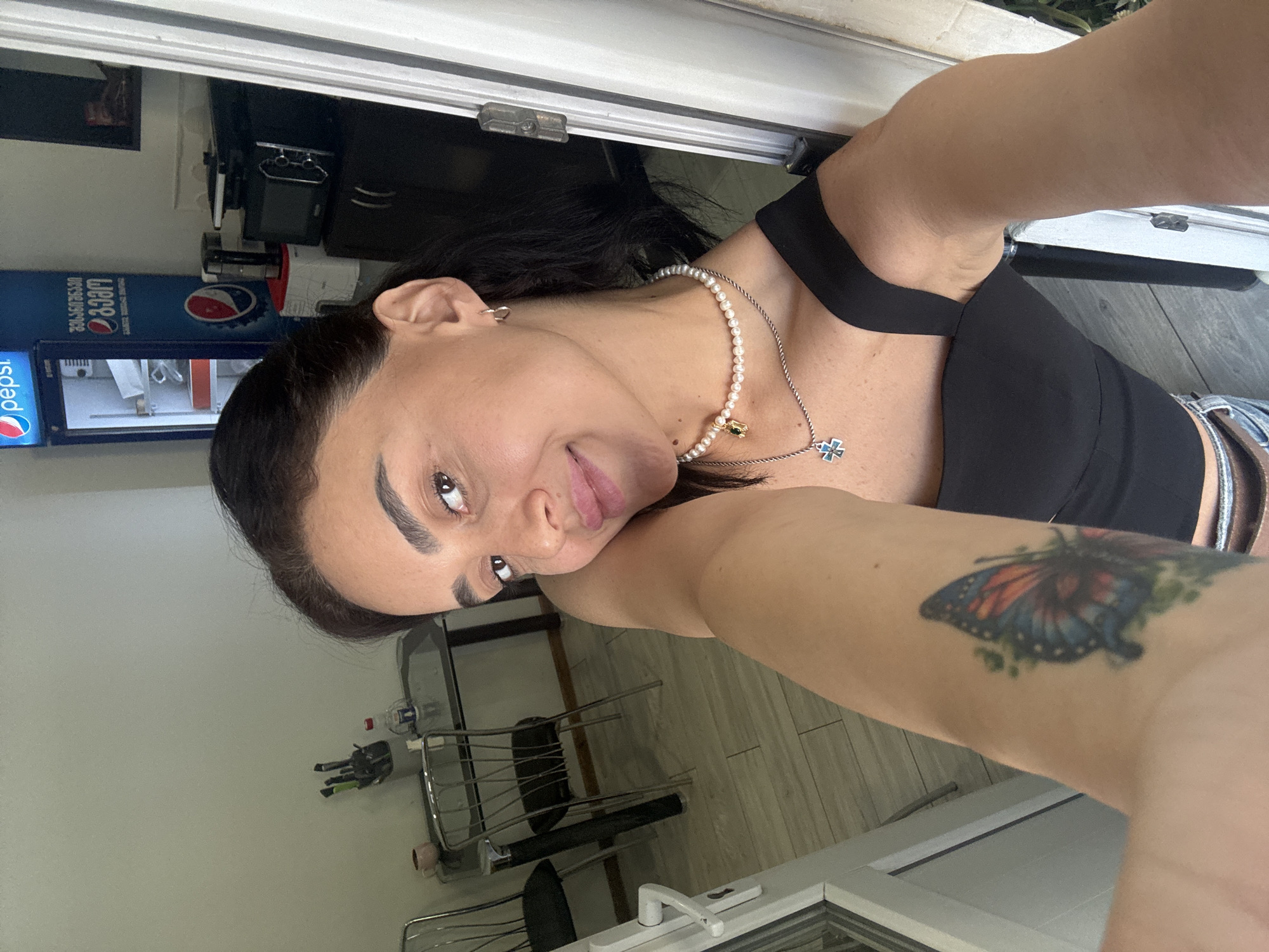 A woman is taking a selfie in what appears to be an interior space that has elements of a kitchen or dining area, with a dining table and chairs visible in the background. A Pepsi branded refrigerator and a microwave are also present. The woman is wearing a black top and has a butterfly tattoo on her left arm. She is also wearing a pearl necklace with a pendant. The setting appears to be residential, possibly a home or a small business. The time of day is not discernible from the image.