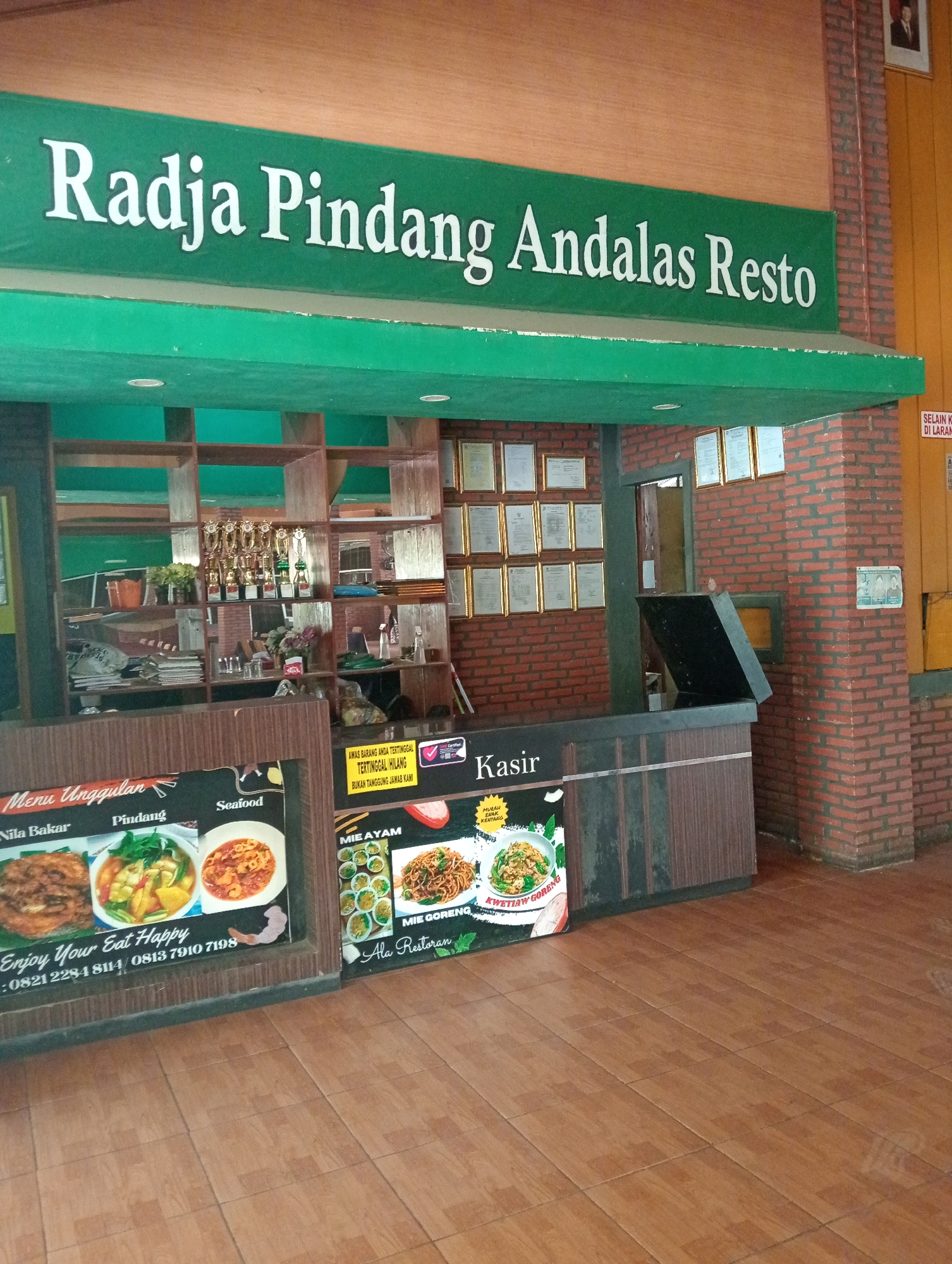 This image shows the exterior of a restaurant named "Radja Pindang Andalas Resto". The restaurant has a green awning with the name of the restaurant written in white, stylized lettering. Below the awning, there is a counter area with menus displayed. One menu shows various dishes including "Nila Bakar", "Pindang", and "Seafood". Another menu displays "Mie Ayam" and "Kwitiaw Goreng". The restaurant appears to be located in Tambak Rejo, Indonesia, as indicated by a sign on the menu with phone numbers. The interior of the restaurant shows shelves with trophies, decorative items, and what appears to be a cash register. The building has a brick facade and tiled flooring. There are no people visible in the image.
