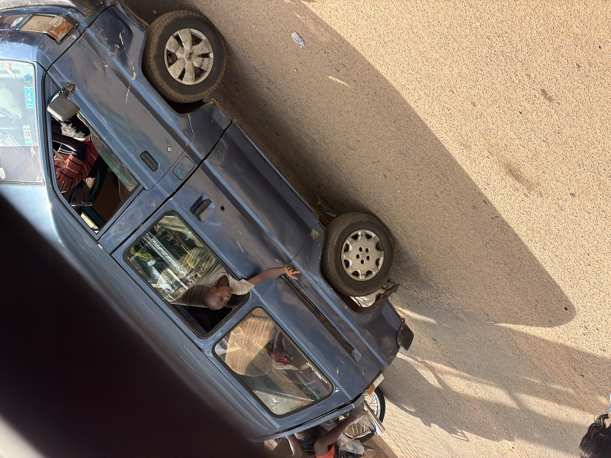 An over-the-shoulder, wide shot captures a dusty, unpaved street scene in Bukuru, Nigeria. A blue, boxy minivan, tilted at an unnatural angle, dominates the foreground. Sunlight casts long, sharp shadows across the ochre-colored ground. A young man with a shaved head, wearing a light-colored shirt, leans out of the minivan's open passenger window, his right arm resting on the frame. His gaze is directed forward, and his mouth appears slightly open. Inside the minivan, a driver can be seen wearing a red and white striped shirt. To the left of the minivan, a portion of a motorcycle is visible, with a person wearing orange shorts and a dark shirt seated on it. The minivan exhibits some visible wear and tear, including scuffs on its side. The scene is devoid of any discernible signage or specific landmarks that would further pinpoint the exact location within Bukuru.