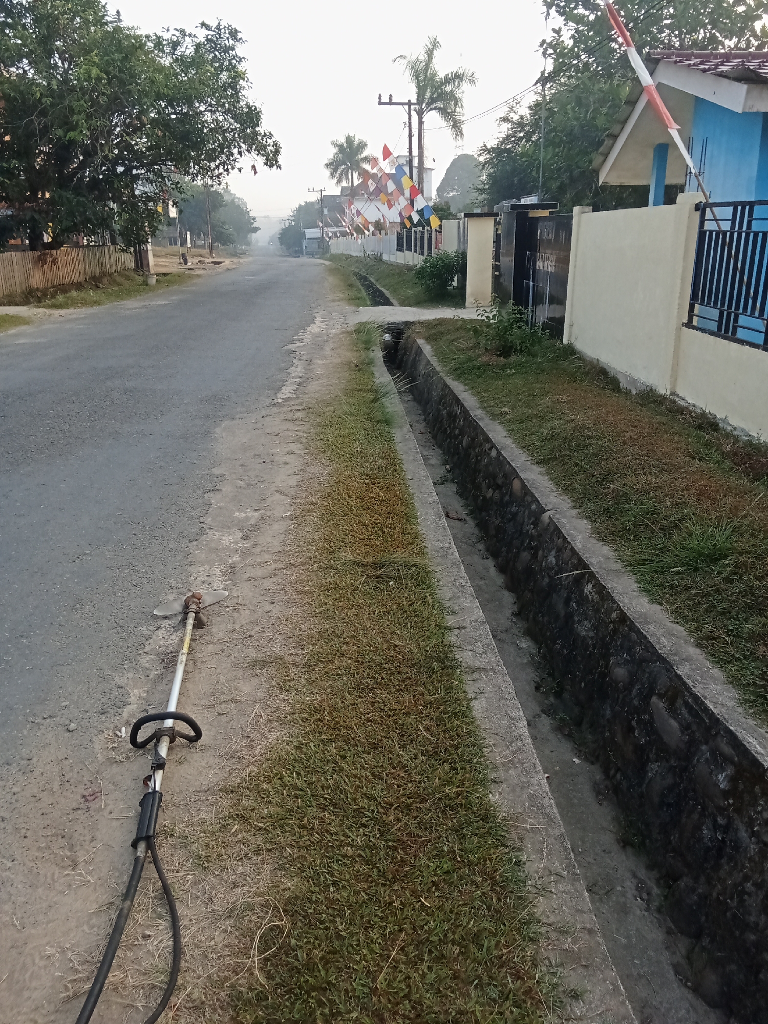 The image depicts a quiet, rural street scene in the village of Tebing Bulang, Indonesia, at what appears to be early morning or late afternoon due to the soft lighting. A paved road runs through the center of the frame, flanked by a grassy embankment and a concrete drainage ditch on the right side. A lawnmower with a metal shaft and a black handle lies on the dirt shoulder of the road in the foreground, suggesting recent or imminent use.
To the right, a light blue house with a red-tiled roof stands behind a low, cream-colored wall and a dark metal fence. A series of colorful pennants, some bearing the Indonesian red and white colors, are strung along the fence and across the road in the background, possibly indicating a local celebration or event. Trees with dense foliage line the left side of the road, with a wooden fence visible behind them. The overall atmosphere is peaceful and suggests a residential area on the outskirts of a community.