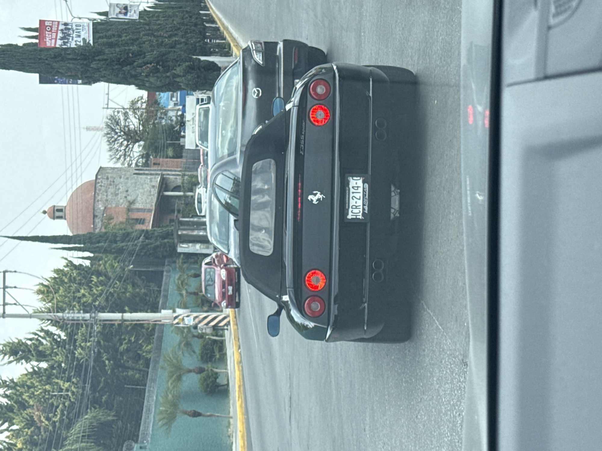 The image shows the rear of a black Ferrari 355 Spider in traffic in what appears to be Puebla, Mexico, based on the license plate, which reads "HCR-214-C" with the words "LA RECTA" below. The car is positioned in a traffic lane, likely on a multi-lane road. A gray Mazda is directly in front of the Ferrari. Other cars are visible further ahead. The environment includes lush green trees and foliage along the roadside. In the background, there is a building with a dome. A billboard is visible as well. The image was taken from inside another vehicle.