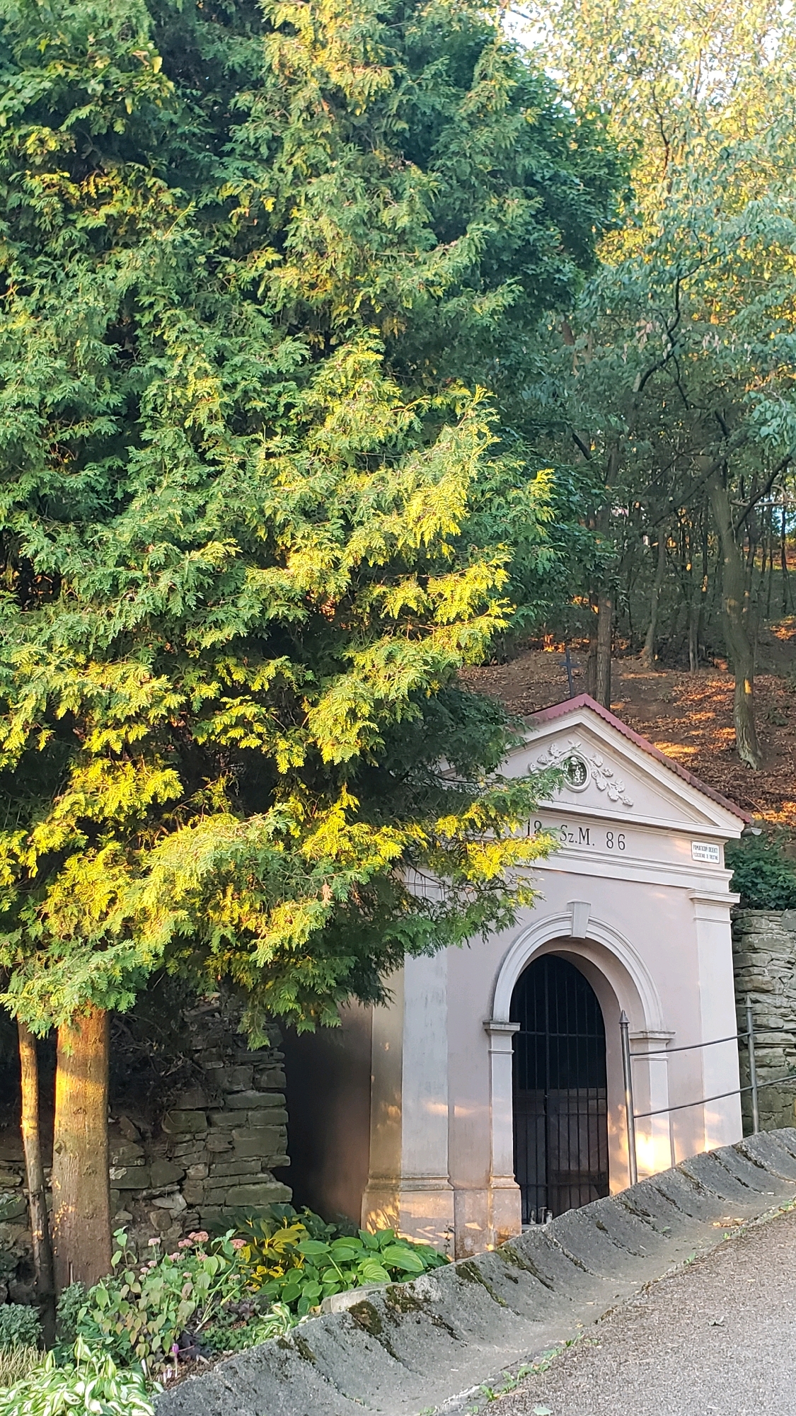 This is a photograph of a small, light-colored building with an arched entrance and a pitched roof, situated in a wooded area with a stone wall and foliage. Above the arch, the building is adorned with decorative carvings and the inscription "18 SZ.M. 86." A dark metal gate is visible behind the arch. The building appears to be a historical structure, possibly a chapel or a crypt. The surrounding environment is natural, with trees, fallen leaves, and a cobblestone path. The lighting suggests it is late afternoon or early morning, with sunlight filtering through the trees and casting warm hues on the scene. This structure is located in the cemetery of the Church of St. Nicholas in Prešov, Slovakia.