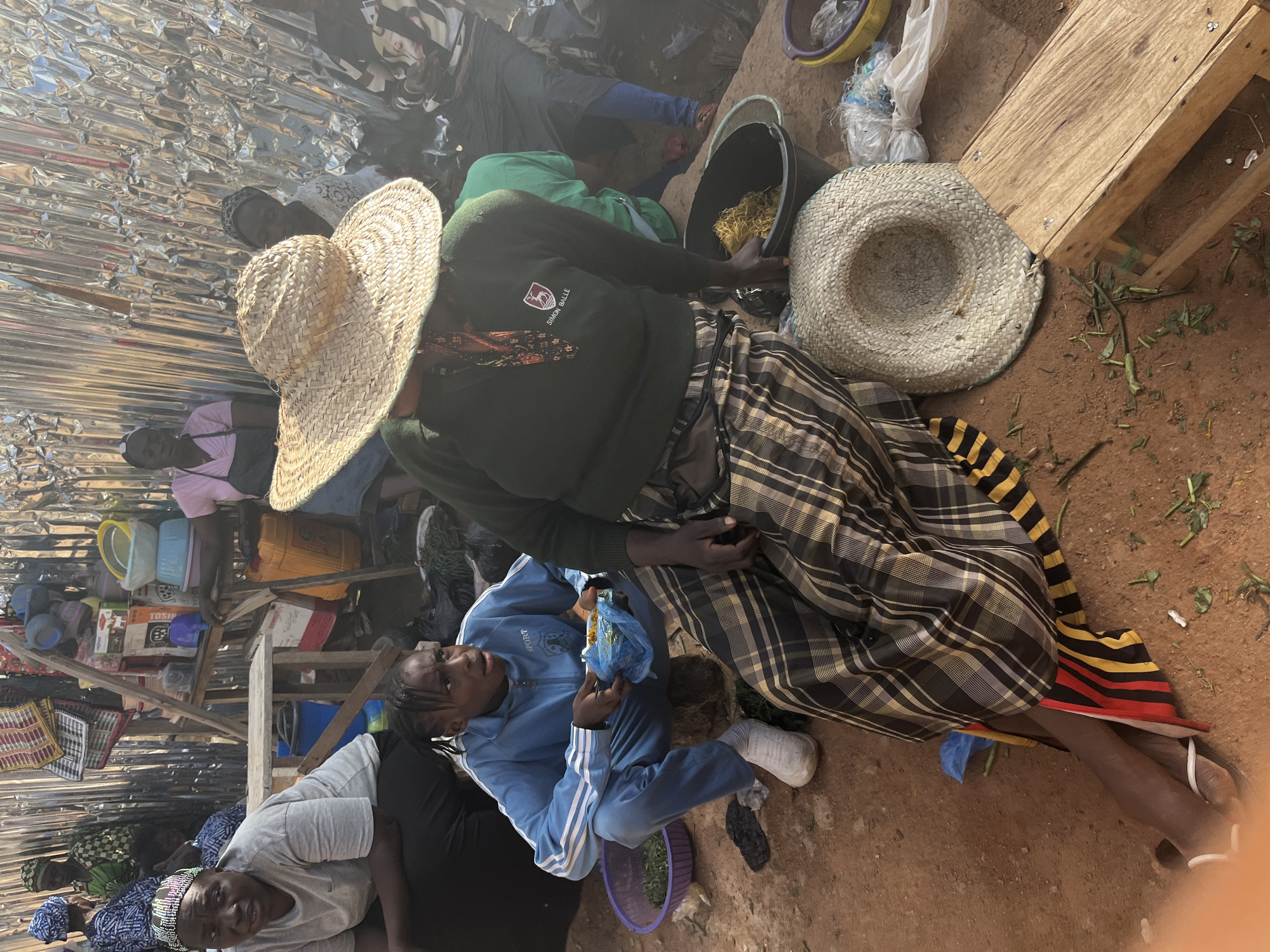 The image depicts a bustling market scene, likely in Jos, Nigeria, characterized by a vibrant, outdoor setting. In the foreground, a young person in a blue tracksuit is seated, holding a plastic bag with food. Beside them, an adult wearing a straw hat and a patterned skirt is also seated, interacting with the market environment. The background reveals stalls filled with various goods, including containers, baskets, and what appear to be food items. Several other individuals are visible in the background, engaged in market activities. The ground is a reddish-brown dirt, indicative of an outdoor market. The overall atmosphere suggests a lively commercial hub with a strong sense of community.