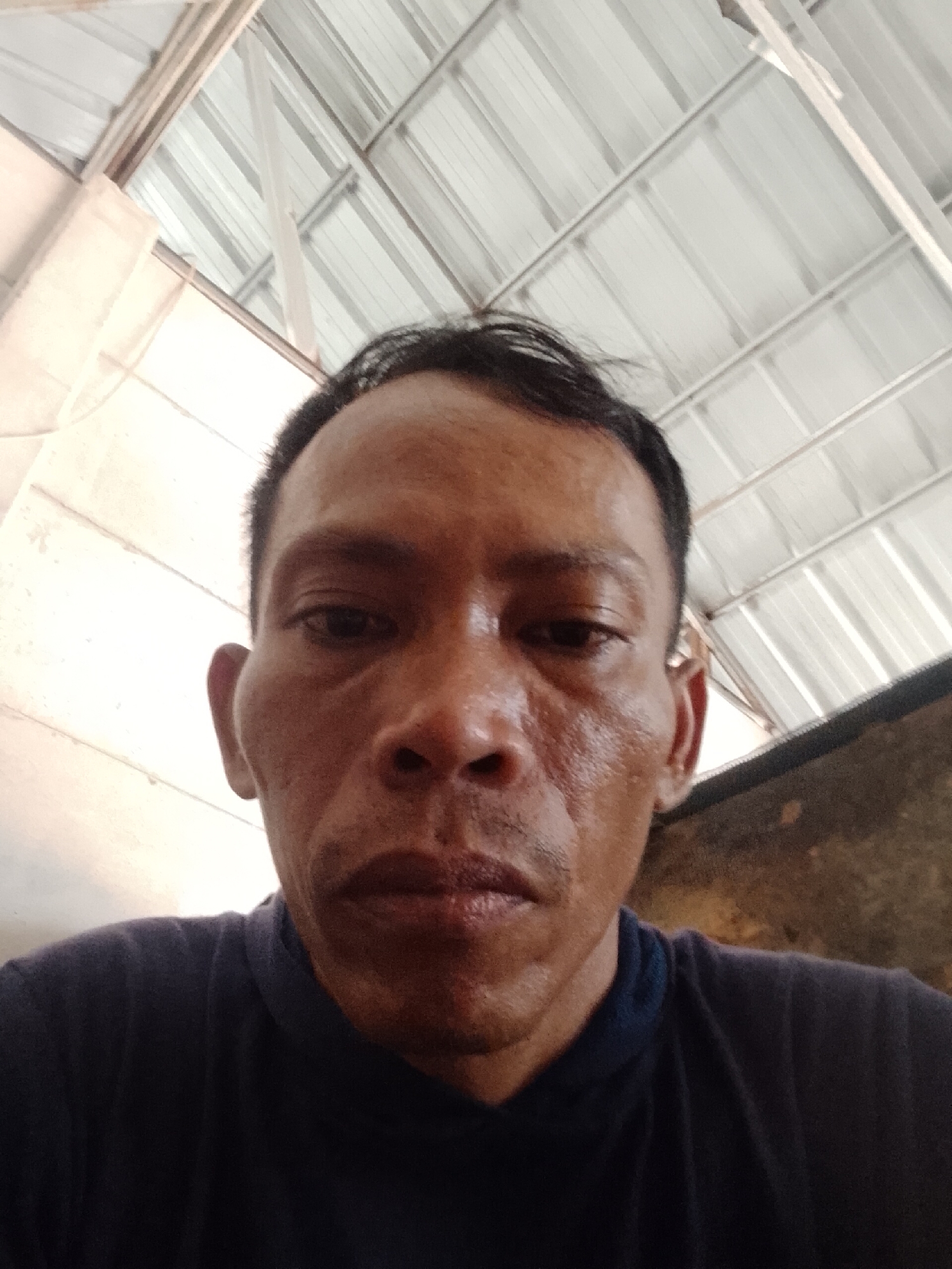 A close-up shot of a man with dark skin and short black hair, looking directly at the camera. He is wearing a dark blue shirt. The background shows a corrugated metal roof, suggesting an indoor or partially covered outdoor space. Based on the location information provided, the photo was taken in Bukit Ketok, Indonesia. The man's face appears sweaty, and his expression is neutral. The lighting is somewhat diffused, illuminating the face from above. A dark shadowy surface is visible to the right of the man's head.