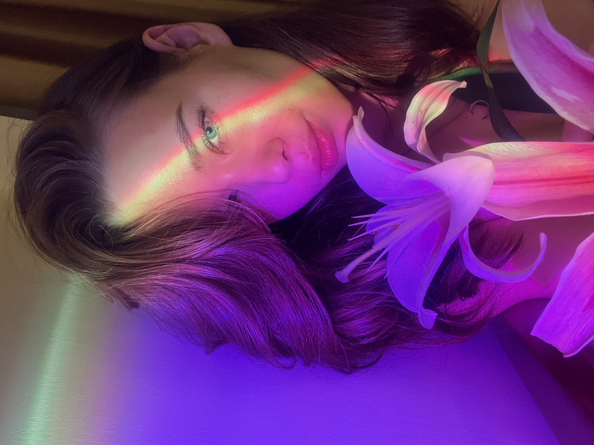 A medium close-up, diagonal shot features a person, likely female, with long, dark brown hair and light green eyes, looking directly at the viewer. The person's face is partially illuminated by a striking rainbow light streak across the forehead and left eye, casting a vibrant glow. A soft smile is present on her lips.
The scene is awash in rich, artificial lighting, primarily dominated by purples and pinks, especially noticeable on the lower half of the image, the person's hair, and the surrounding elements. Several large, pink and white lily flowers are arranged around the person's head and hair, with their petals catching the colorful light. The person's hair cascades around the face and flowers, exhibiting purple highlights from the ambient lighting.
The overall mood is artistic and serene. There are no visible location cues within the image itself that suggest the Nam Natural Heritage Area Trang, Vietnam; the lighting appears to be controlled and artificial, possibly for a photoshoot in a studio or indoor setting. No text is visible in the image.