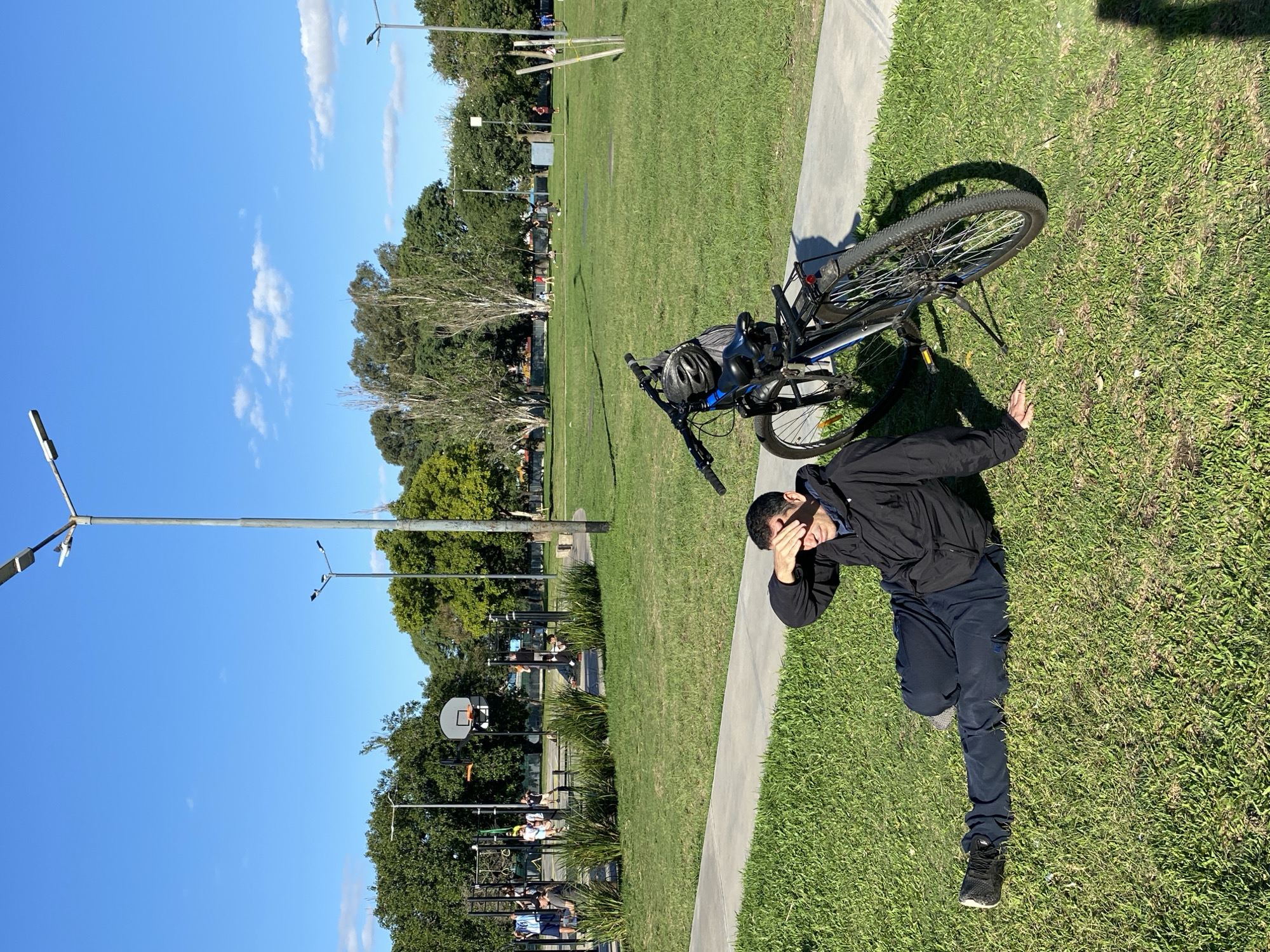 A person is lying on the grass next to a bicycle in a park. The person is wearing a black jacket and dark pants, and is covering their face with their hand. The bicycle is black and blue. The park has a paved walkway next to the grass. In the background are trees, streetlights, and what appears to be a basketball court. The location appears to be outdoors in a park setting, but I cannot determine if this image was taken in Buenos Aires, Argentina.