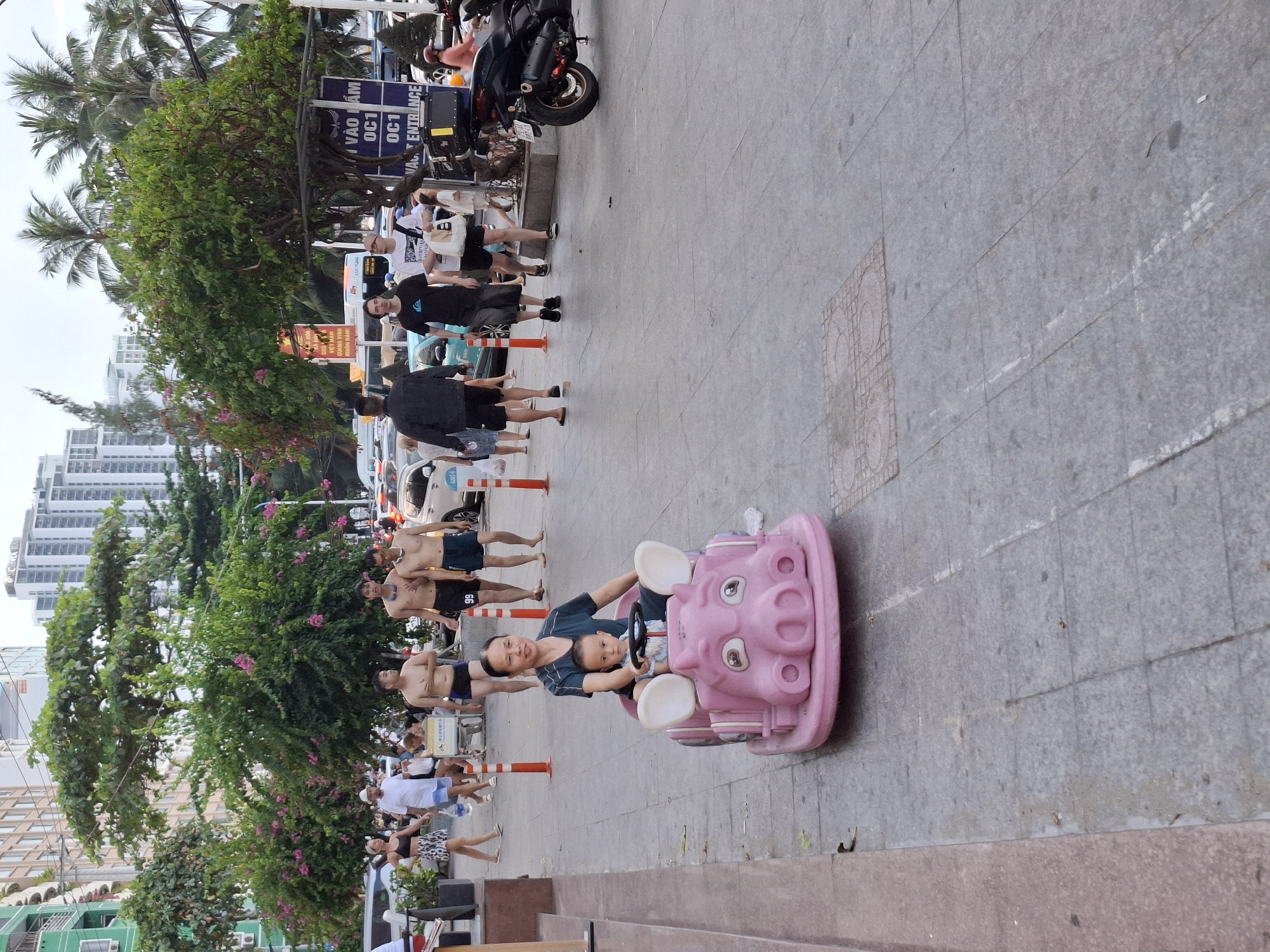 A woman and a young child are seated in a pink pig-shaped toy car on a paved sidewalk. The woman is in the driver's seat, and the child is in the passenger seat. Both are facing forward. The woman is wearing a dark blue top, and the child is wearing a light blue and white striped shirt. The toy car is positioned on the left side of the frame, with its front facing towards the bottom right.
In the background, several other people are walking on the sidewalk. To the left of the toy car, there are orange bollards and the edge of a building. Further in the background, there are trees, palm trees, and tall buildings, indicative of a city environment. Traffic, including cars and motorcycles, is visible on a street beyond the sidewalk. A sign with Vietnamese text is also present in the background. The overall scene appears to be an outdoor public space in a city, likely a pedestrian area near a street.