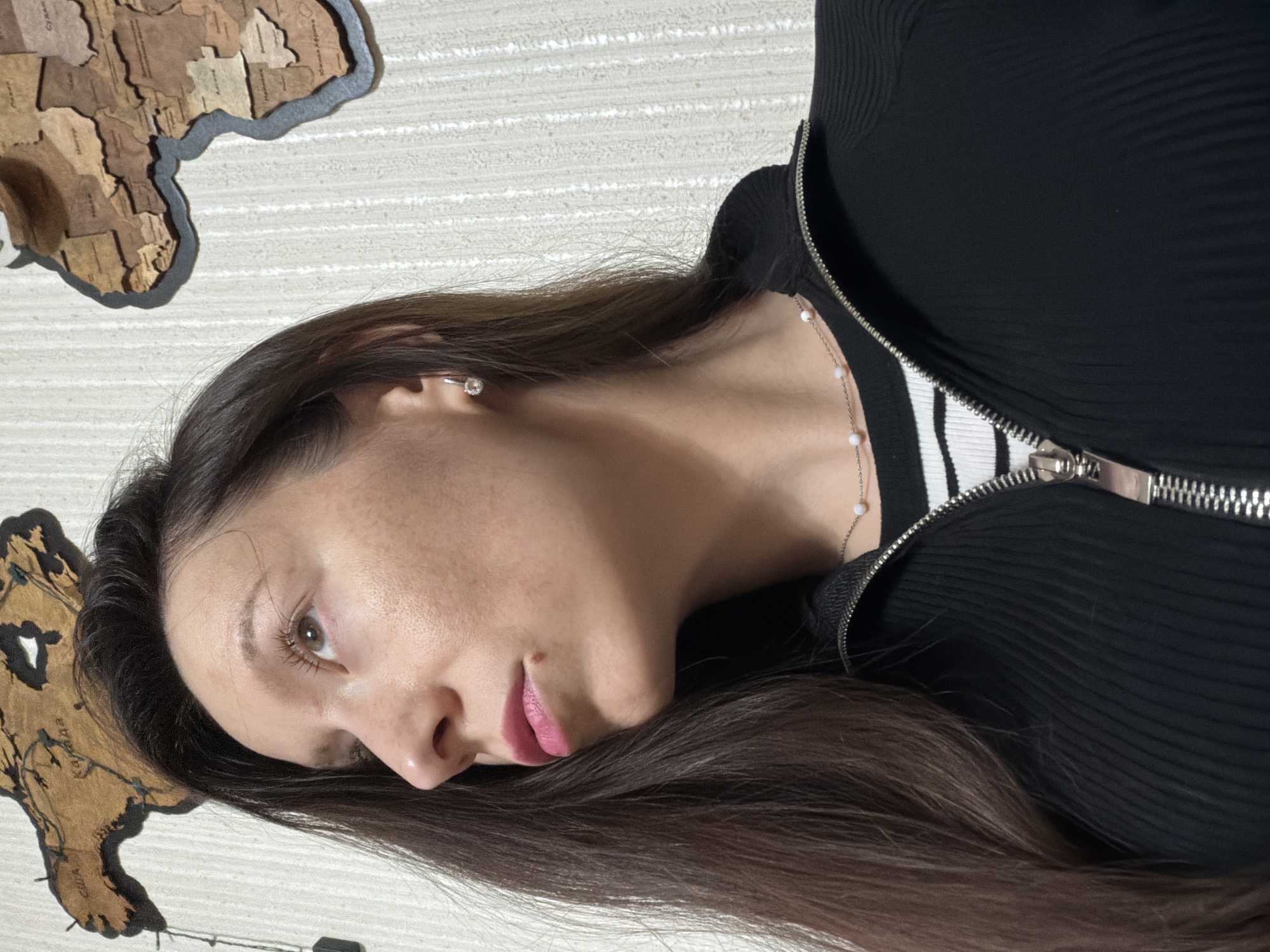 A close-up, profile shot focuses on the face and upper torso of a woman with long, dark brown hair. She is looking upwards and to her left, with her head slightly tilted. Her makeup includes pink lipstick and a small beauty mark visible on her chin. She is wearing a black zip-up jacket over a striped shirt and a delicate silver necklace with small pearls. She has a single stud earring in her visible ear. The background is a textured light-colored wall with two decorative wooden world map pieces mounted on it; one depicting North America and the other Africa. The scene appears to be indoors, possibly in a home. The location is identified as Raevka, Russia.