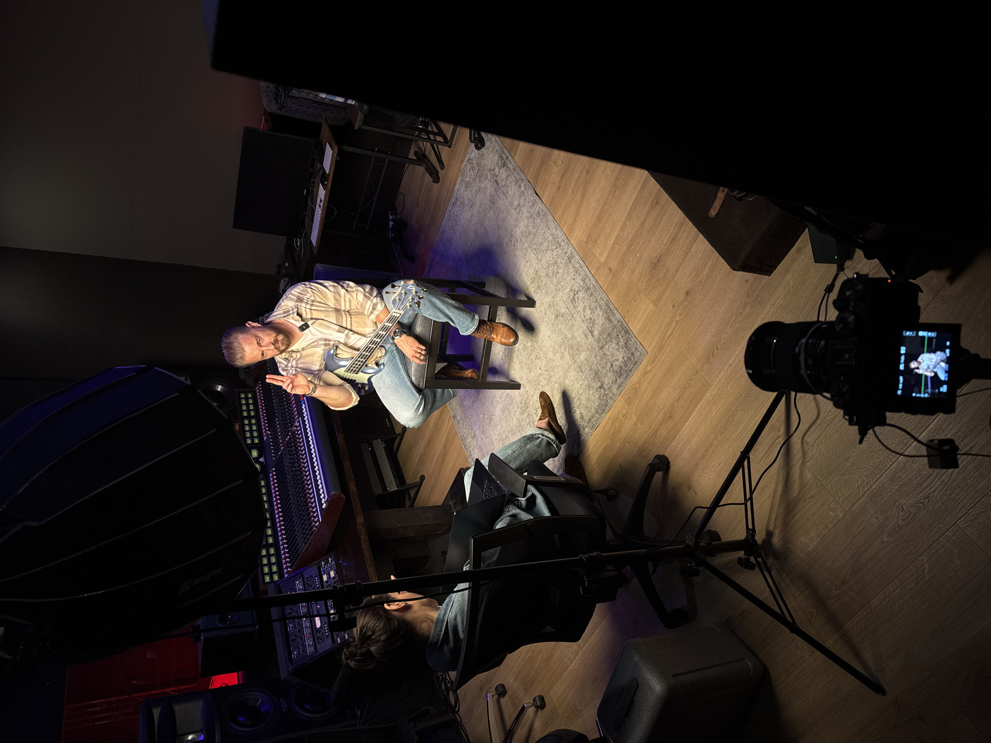 The image captures a scene in what appears to be a recording studio or creative workspace, potentially located in Santa Monica, California, United States, judging by the interior design style and potential clientele associated with such facilities in that city.
A man is seated on a stool, holding and apparently playing an electric guitar. He is wearing jeans and a checkered shirt. He is positioned near a large audio mixing console. A woman is sitting in a rolling chair, facing away from the camera. She appears to be operating a laptop, possibly directing or monitoring the recording session.
A professional camera mounted on a tripod is pointed towards the man with the guitar. A large softbox lighting setup is positioned on the left side of the frame, providing illumination for the scene.
The room has wood flooring, with a rug placed centrally under the area where the man is seated. Audio equipment, monitors, and various studio-related items are visible in the background. The lighting suggests a focused, controlled environment optimized for recording.