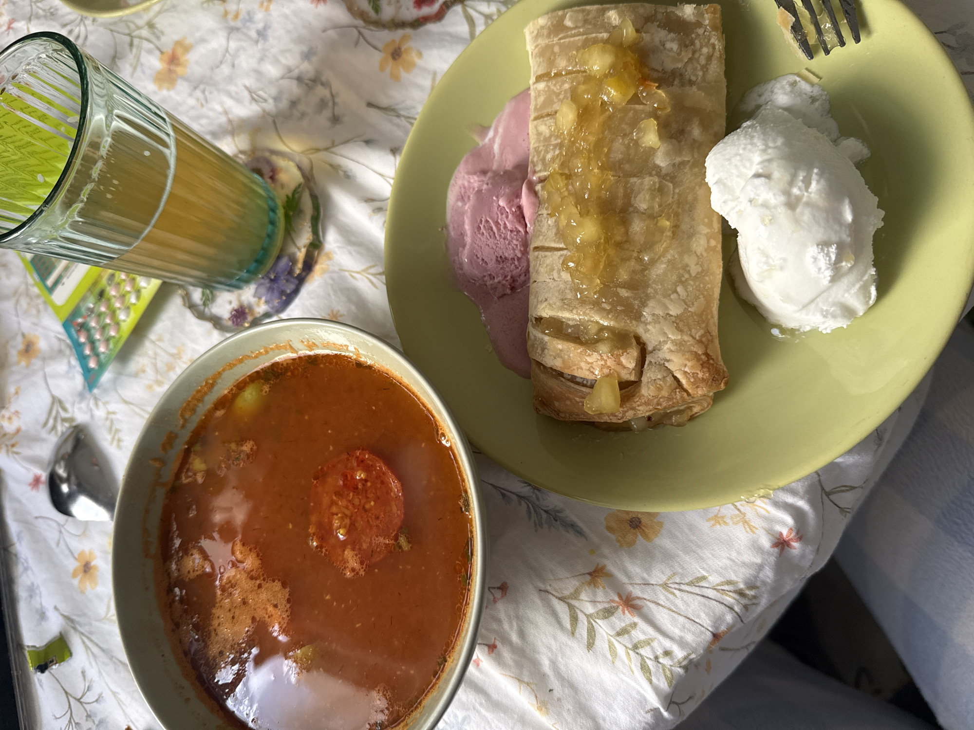An overhead shot displays a diverse meal arranged on a white floral tablecloth or bedspread, suggesting a relaxed, indoor setting, likely in the City of Zagreb, Croatia.
The central subjects include a bowl of reddish-orange soup, possibly tomato or lentil, featuring a visible slice of tomato and some herbs, accompanied by a small spoon. To the right, a light green plate holds a golden-brown pastry, resembling a strudel, topped with a chunky, glistening fruit filling, likely apple. Beside the pastry are two scoops of melting ice cream: one pink or berry-flavored, and another white, possibly vanilla. A metal fork is partially visible near the white ice cream.
To the left, a tall, fluted glass contains a light yellow or orange beverage, resting on a decorative coaster with purple and green floral patterns. Next to the glass, a small, rectangular item, possibly a blister pack for medication or candy, with a grid of small circles, is visible. A light blue fabric, likely a blanket or clothing, is seen on the far right. The soft lighting indicates daytime. No people or legible text are visible in the image.