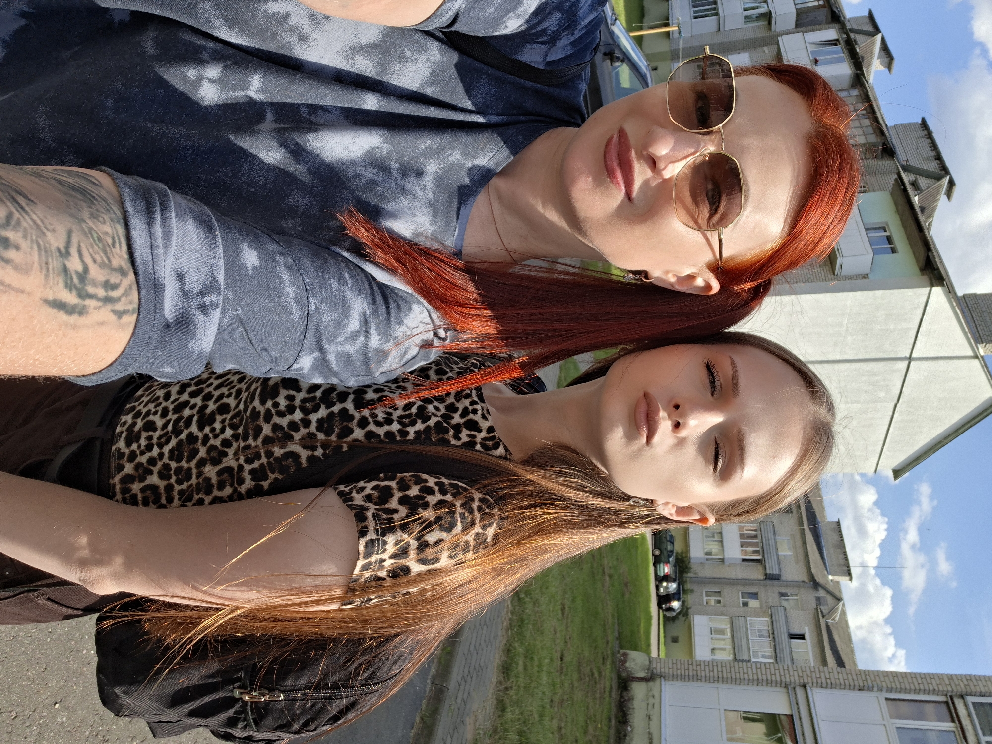This is a selfie taken outdoors, featuring two women standing side-by-side. The woman on the left, with bright red hair styled in pigtails and wearing large, hexagonal sunglasses and a tie-dye t-shirt, is in the foreground and closer to the camera. The woman on the right, with long, auburn hair and wearing a leopard-print shirt, is slightly behind and to the right of the first woman, making a duckface. In the background, there are multi-story apartment buildings with balconies, a paved walkway, and a grassy area with parked cars. The sky is blue with scattered white clouds. The scene is set in a typical urban residential area, likely in the city of Myadzyel, Belarus, given the architectural style of the buildings.