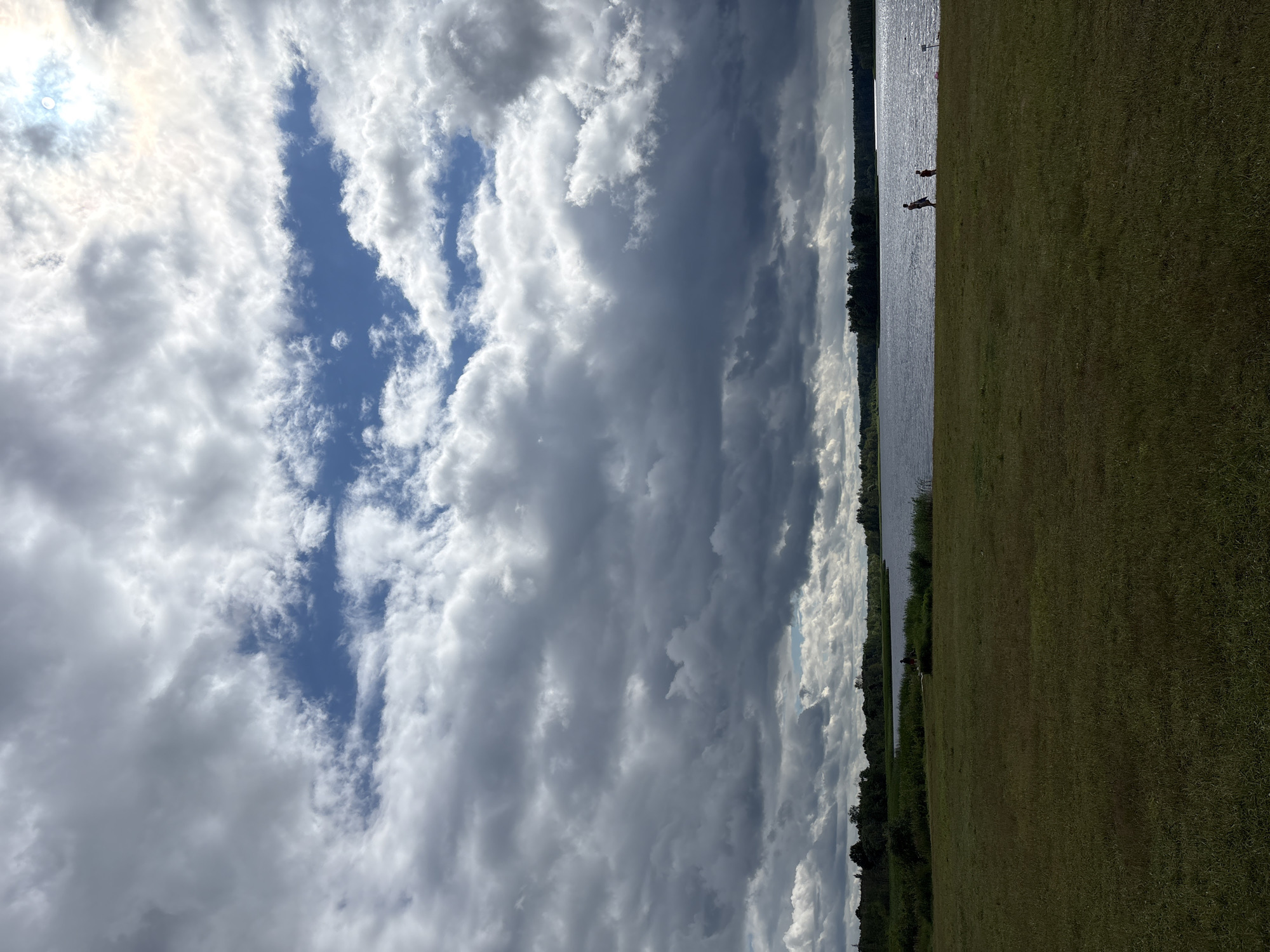 The image shows a wide, grassy field in front of a large body of water. A treeline borders the far side of the water. The sky is dramatic, with large, dark clouds mixed with patches of blue sky and bright sunlight. Two silhouetted figures, one adult and one child, are walking near the water's edge in the middle ground. The scene is likely an outdoor recreational area in Мазалаўскі сельскі Савет, Belarus.