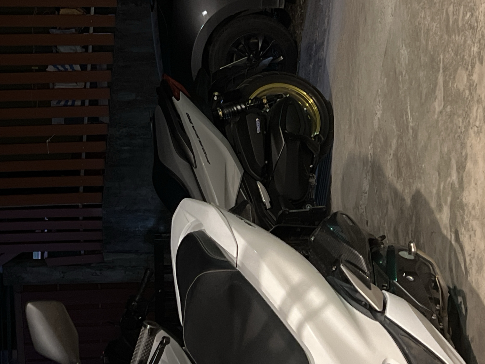 The image shows two motorcycles parked in an outdoor setting at night, with a fence in the background. The motorcycle in the foreground is white with black accents and appears to be a scooter. The second motorcycle, partially visible behind the first, is also a scooter, white with black detailing and a gold-colored rim on its wheel. Next to the motorcycles, a dark-colored car is parked. The ground is concrete. The fence is made of vertical wooden slats. The lighting suggests it is nighttime, with artificial light sources casting shadows. There are no people visible in the image.