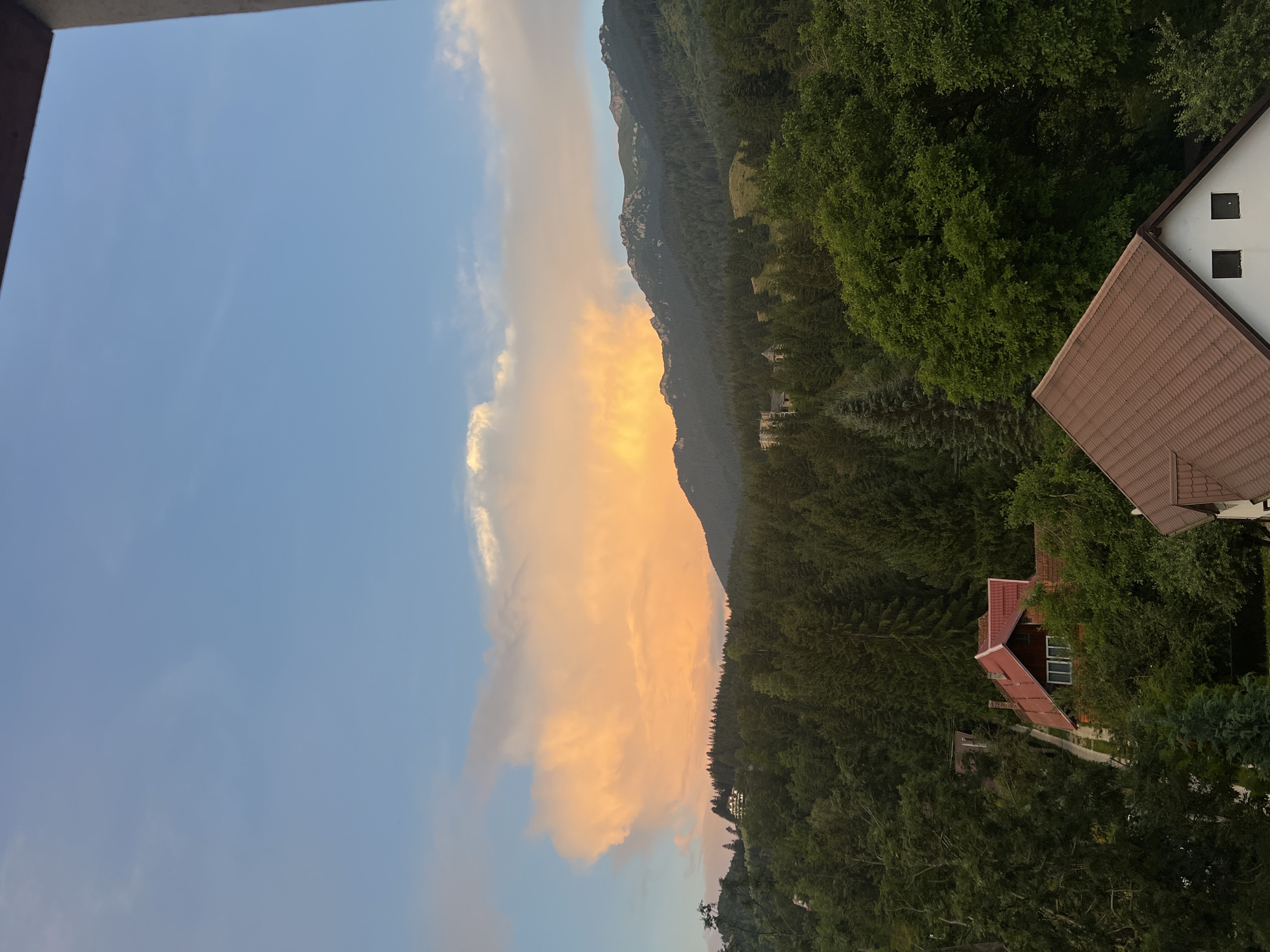 The image shows a view from a balcony looking out at a mountainous landscape. In the foreground, there are the roofs of two houses, one with a red tiled roof and the other with a brown tiled roof. Beyond the houses, a dense forest of evergreen trees covers the lower slopes of the mountains. The mountains themselves are covered in trees as well, with rocky peaks visible at the top. A large, dramatic cloud formation is visible in the sky above the mountains, illuminated by what appears to be the setting sun, casting an orange and pink glow. The sky is a clear blue with some lighter clouds scattered about. The scene appears to be peaceful and serene.