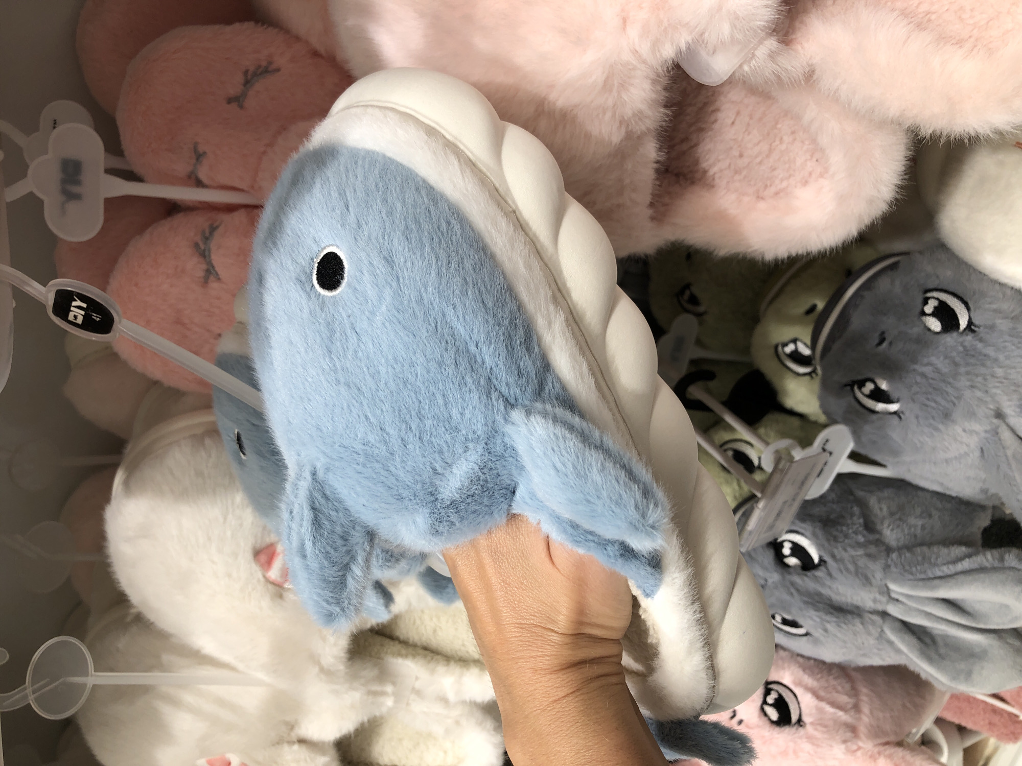This media file shows a close-up of a hand holding a blue plush slipper shaped like a whale. The whale slipper has a white sole and a light blue fuzzy body with a black eye. The background is filled with other plush slippers in various colors, including pink, gray, and white, many of them also designed with animal faces. These slippers are likely on display in a store, possibly a retail shop or market stall, given the hanging tags and the organized arrangement. The lighting suggests an indoor setting, possibly a store with artificial lighting. The overall impression is one of a retail display of soft, whimsical footwear. The image does not provide any cues about the time of day, weather, or specific emotions. There are some visible text elements on tags, such as "DIY" and what appears to be "BIA" on a white tag. The location context provided is Đà Nẵng, Vietnam, which could indicate the origin or retail location of these items.