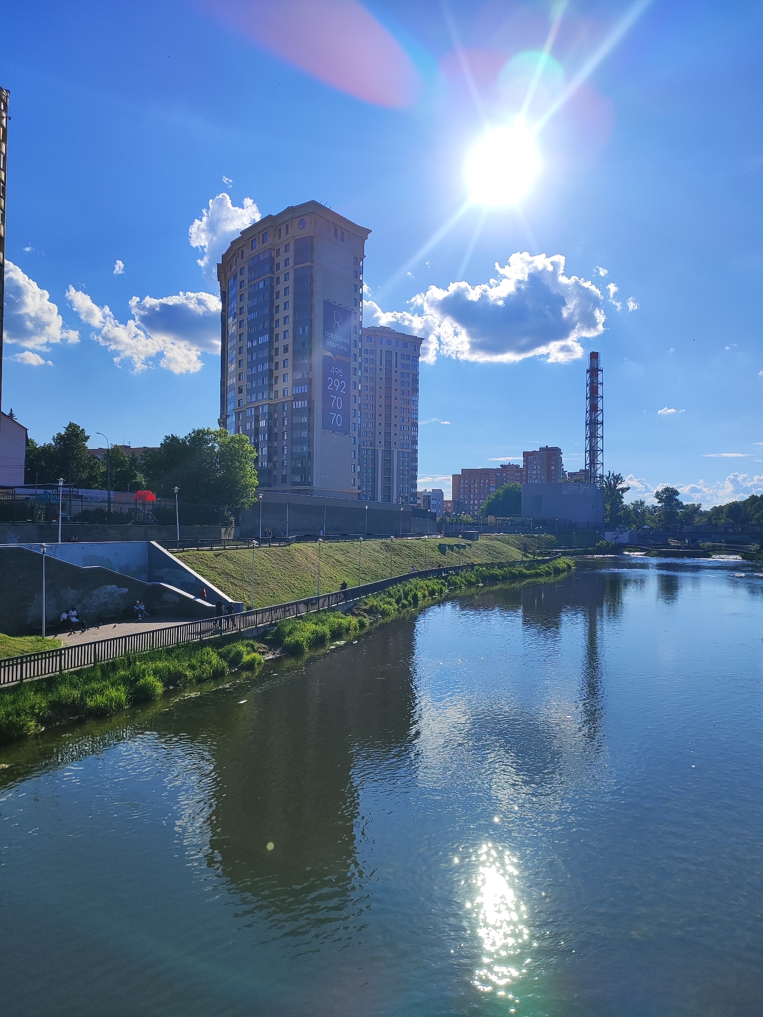This daytime photograph captures a scenic view along a river in an urban setting. The foreground is dominated by the river, its surface reflecting the bright blue sky, scattered white clouds, and the surrounding structures. Sunlight creates a strong lens flare and sparkling reflections on the water, indicating a clear and sunny day, likely late morning or early afternoon given the sun's high position.
On the left bank, a landscaped embankment features green grass and trees, with a paved path running alongside a black metal fence. Several people are visible relaxing or strolling, some seated on concrete steps that descend towards the river. A tall, modern residential building, multi-storied with numerous windows, stands prominently on the left bank. On its facade, a large banner displays Russian text: "ПОСЛЕДНИЕ КВАРТИРЫ" (Last Apartments) and a phone number: "495 292 70 70".
Further down the river on the right, a distinctive red and white striped industrial chimney rises tall, contrasting with the sky. Other urban buildings and structures are visible in the background, including a bridge spanning the river in the distance. The sky is bright blue with scattered, fluffy white clouds. The overall scene depicts a peaceful urban riverfront environment in Shchyolkovo, Russia.