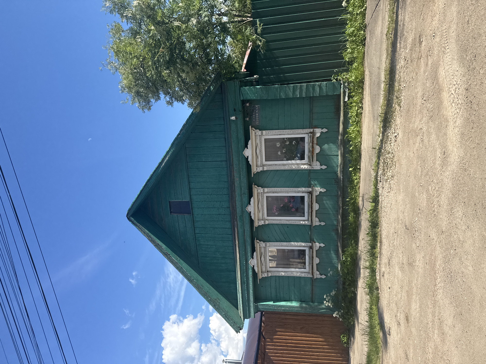 A bright green, wooden, one-story house with a dark green pitched roof sits on a dirt road in Vereya, Russia. The house has three white-trimmed windows with ornate white wooden carvings, and the windows appear to contain potted plants. A sign above the rightmost window reads "УЛИЦА ЛЕНИНСКАЯ" (Leninskaya Street). A green metal fence runs along the right side of the house, and a wooden fence is visible to the left. A large green tree is behind the house and to the right. The sky is blue with scattered white clouds and a sliver of the moon is visible. Power lines are strung across the top of the frame.
