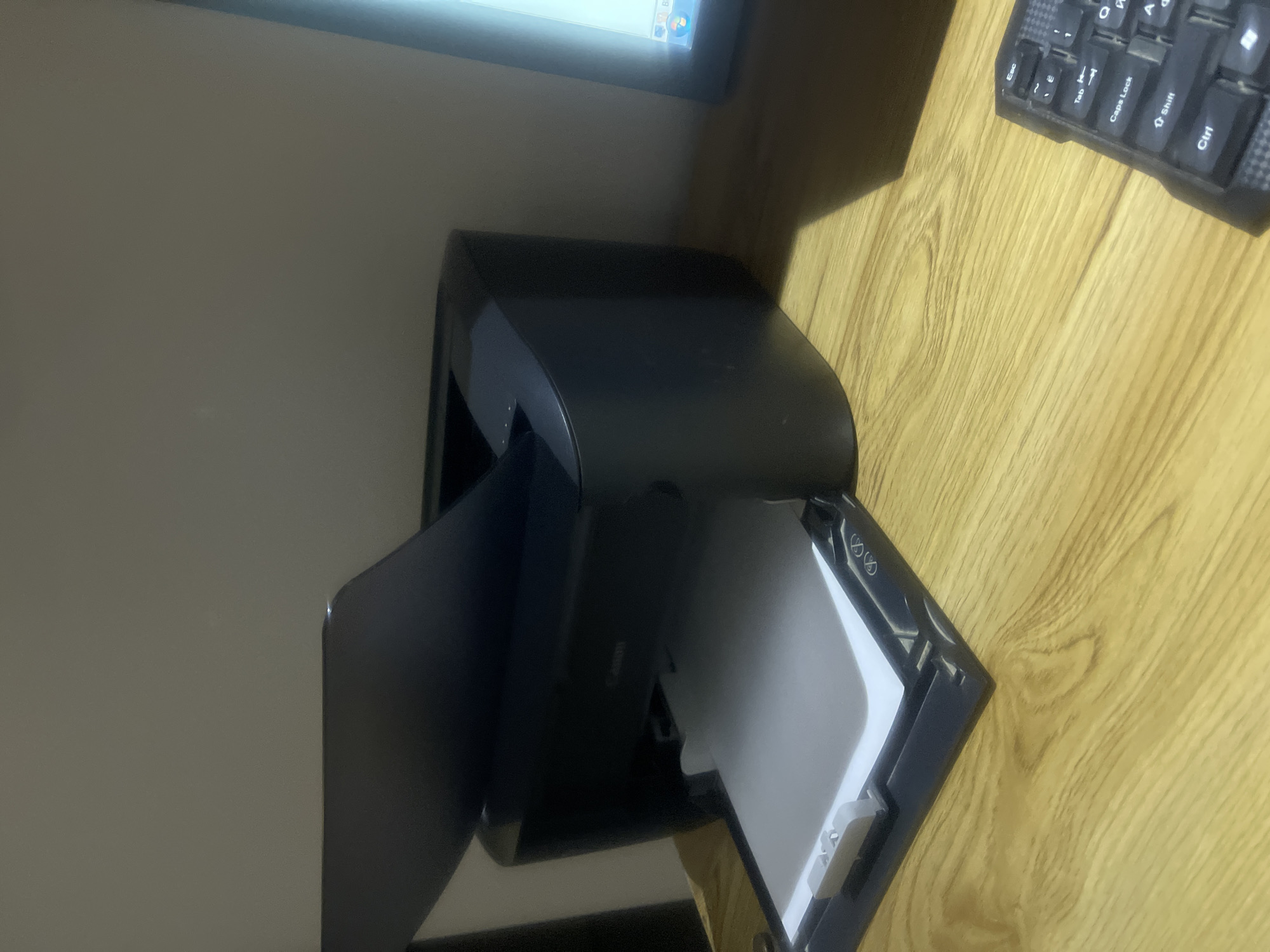 The image depicts a close-up, eye-level, indoor shot of a black Canon printer with a sheet of paper in its output tray. The printer is positioned on a wooden desk, which occupies the foreground and extends into the middle ground. To the right of the printer, a portion of a computer monitor and a black keyboard with visible keys are present. The background consists of a plain, light-colored wall. The lighting appears to be artificial, casting shadows on the desk and wall. The overall scene suggests a home or office workspace. There is no indication of the city of Qarshi, Uzbekistan, in the image.