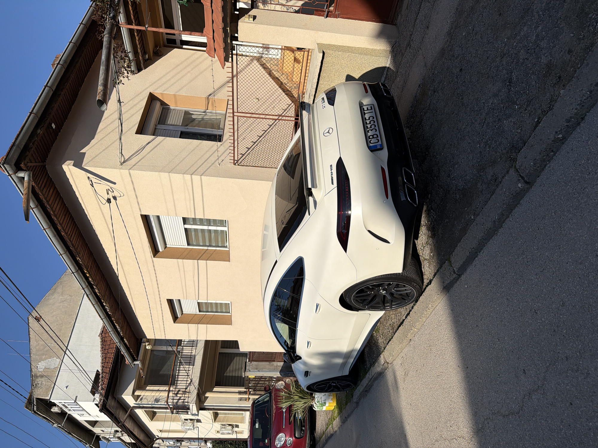 The image shows a white Mercedes-AMG GT 63 S parked on a narrow street in what appears to be a residential area. The car's license plate reads "CB 3555 TE," indicating it is registered in Bulgaria. The background features two-story buildings with stucco facades and metal railings on the balconies. A red car is partially visible behind the Mercedes. Power lines stretch across the sky in the upper left corner of the frame. The setting seems to be Lovech, Bulgaria. The photo was taken during daylight hours, with clear blue skies.
