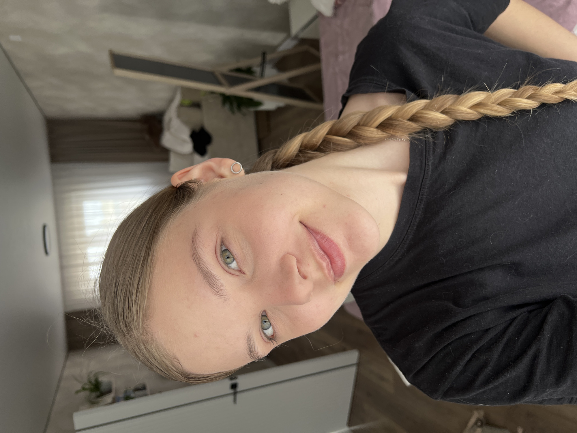 A young, fair-skinned woman with light blonde hair braided down her right side is the primary subject of the image. She is wearing a black t-shirt. The woman is looking towards the camera, with a slight smile. She has green eyes. A small silver hoop earring is visible in her left ear.
The background reveals a partially visible room. Elements include a white dresser, a shelf with various items, and a glimpse of curtains over a window. The image is rotated, such that the ceiling appears on the left and the floor on the right. The location cannot be determined to be Votkinsk, Russia from the information available in the image.