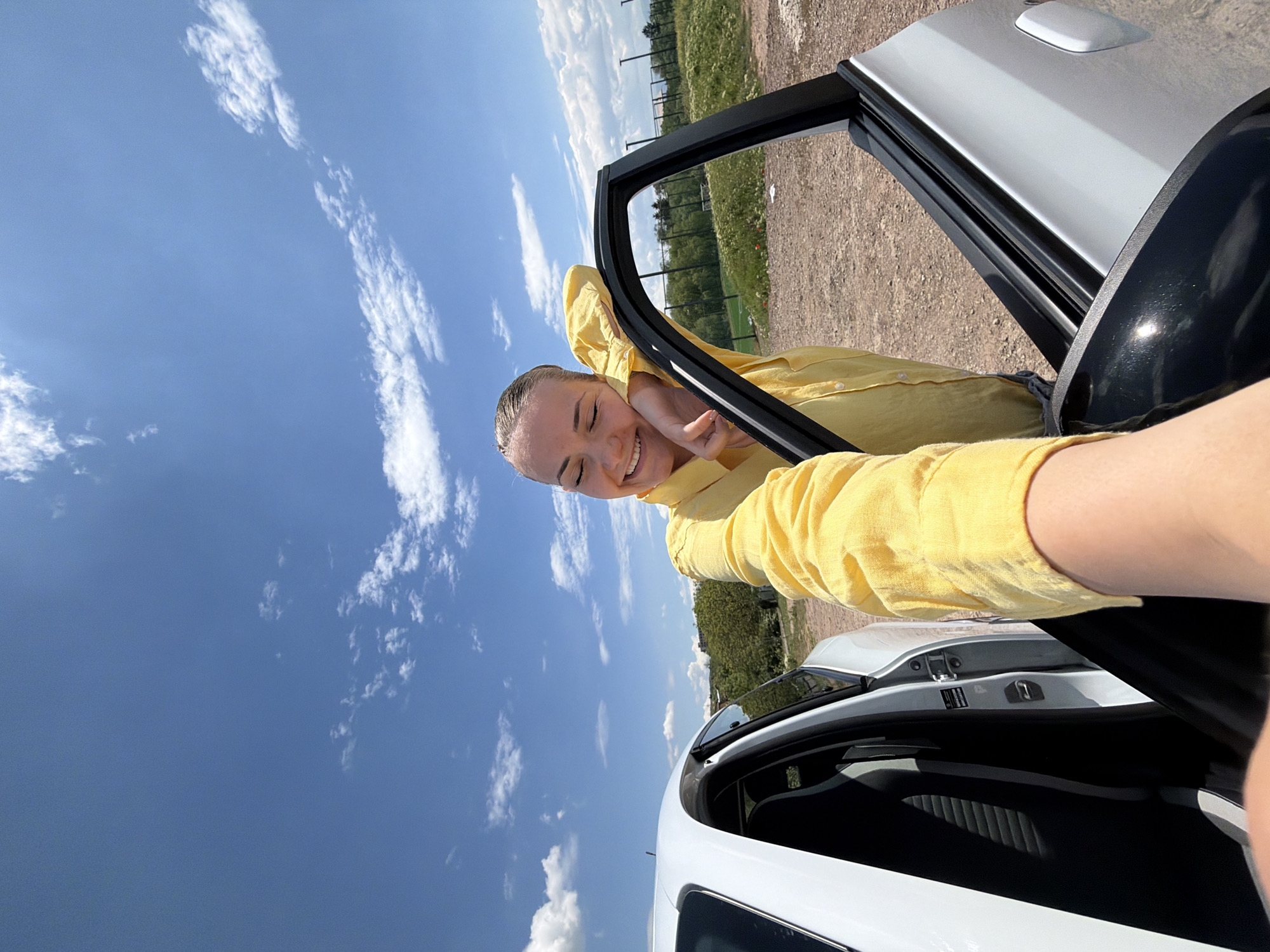 A young woman in a yellow long-sleeved shirt is hanging out the open door of a white car. She is smiling with her eyes closed. The sky is blue with scattered white clouds. In the background, there is a field and some trees. The location is unknown and cannot be determined from the image.