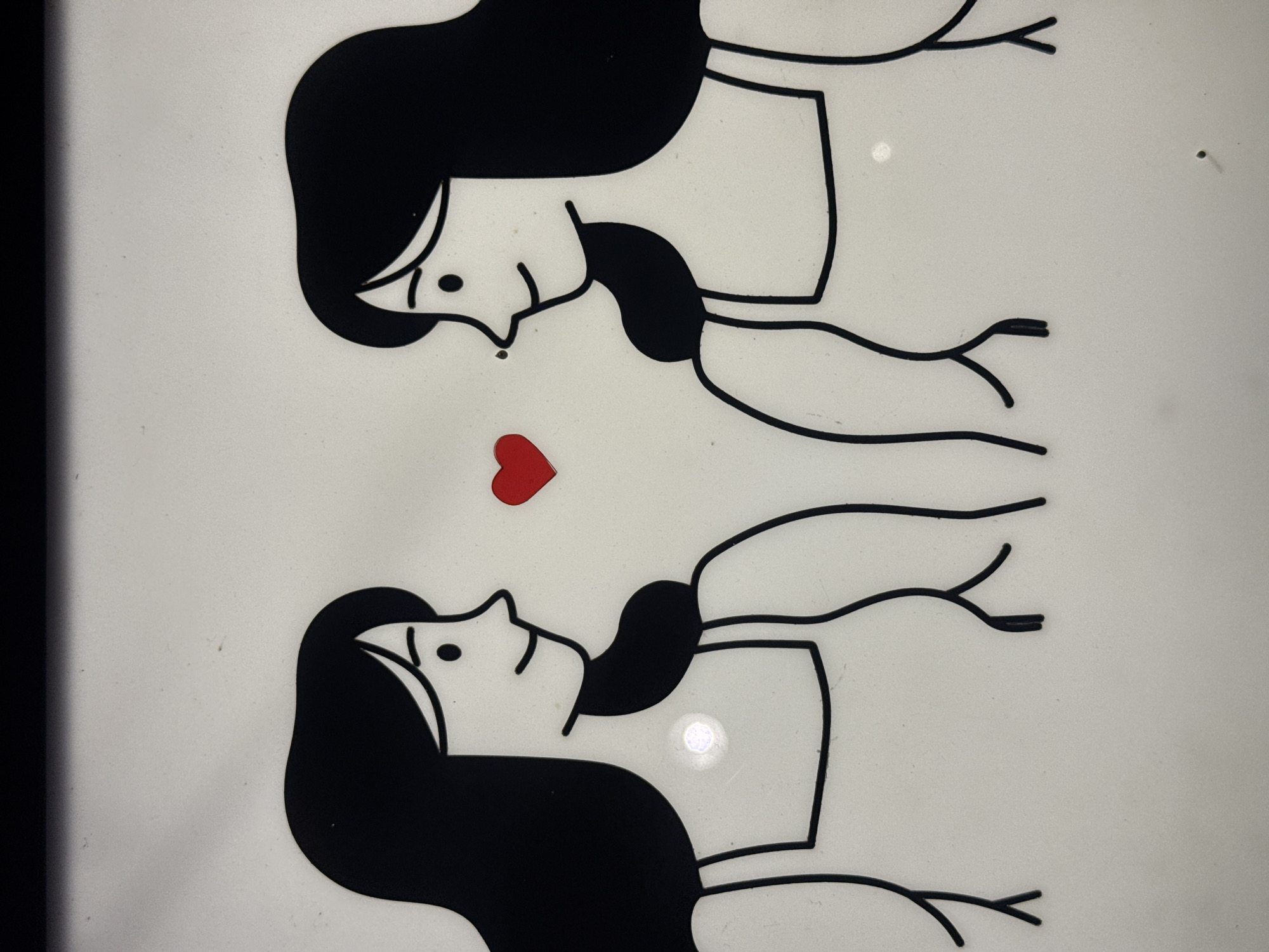 The image displays a minimalist graphic illustration featuring two stylized figures of women facing each other, separated by a small red heart. Both figures have black hair and wear simple white outfits with straps. Their faces are depicted with simple lines for eyes, noses, and smiles. The background is a plain, off-white surface with a black border at the top. There are no discernible details or context that would allow for identification of a specific location within Lâm Viên - Đà Lạt, Vietnam. The image is a static representation, and no actions or interactions beyond the visual suggestion of connection through the heart are present.