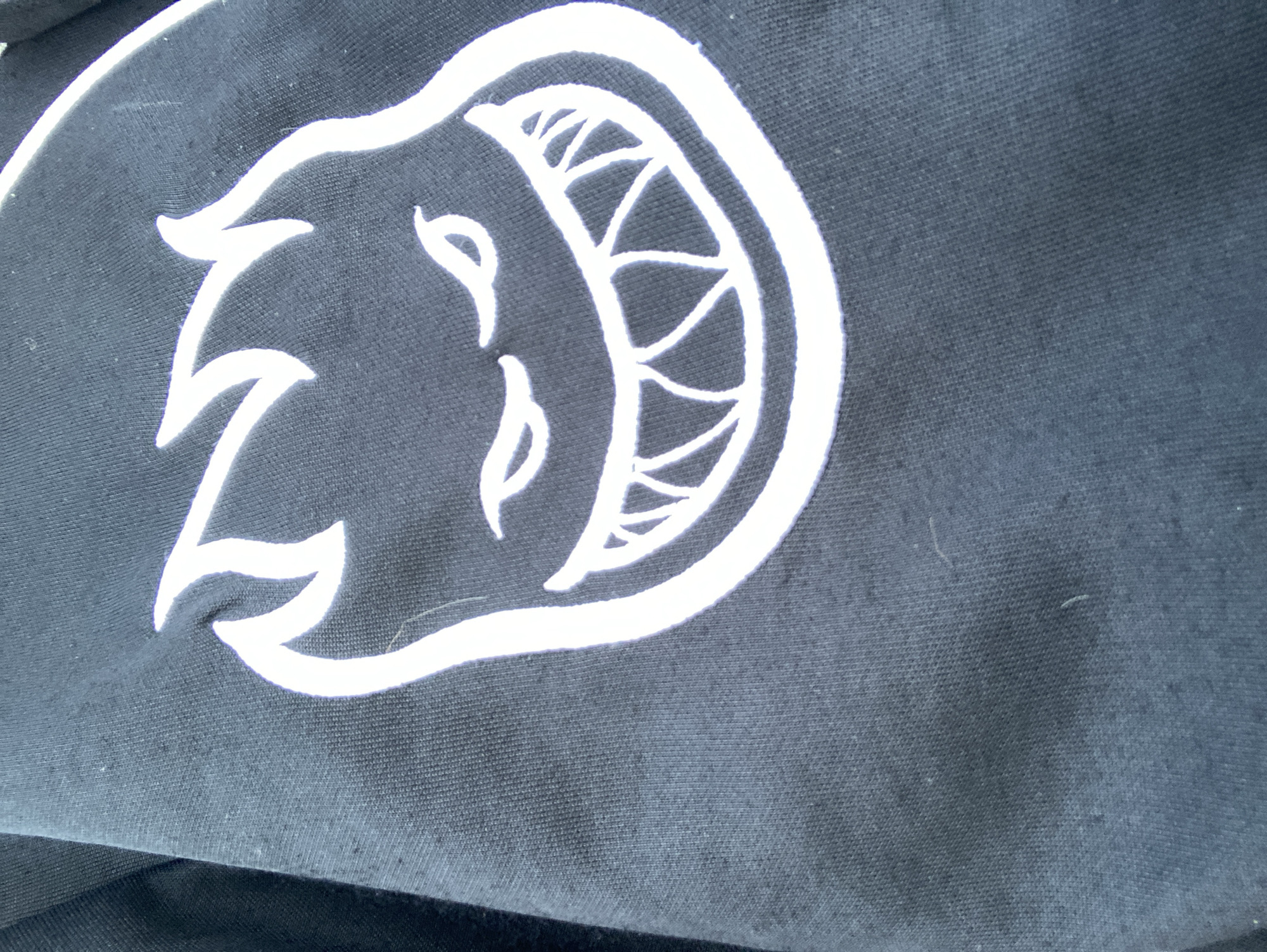 A close-up, high-angle shot shows a white graphic of a cartoonish, fiery-headed creature with a wide, toothy grin printed on a dark fabric, likely clothing. The graphic is centrally located and dominant in the frame. The fabric appears to be textured, possibly cotton, and exhibits some minor wear or lint. The lighting suggests an outdoor setting, though no specific environmental details are visible. There are no people, animals, or any indications of a city location, including Barcelona, Spain.