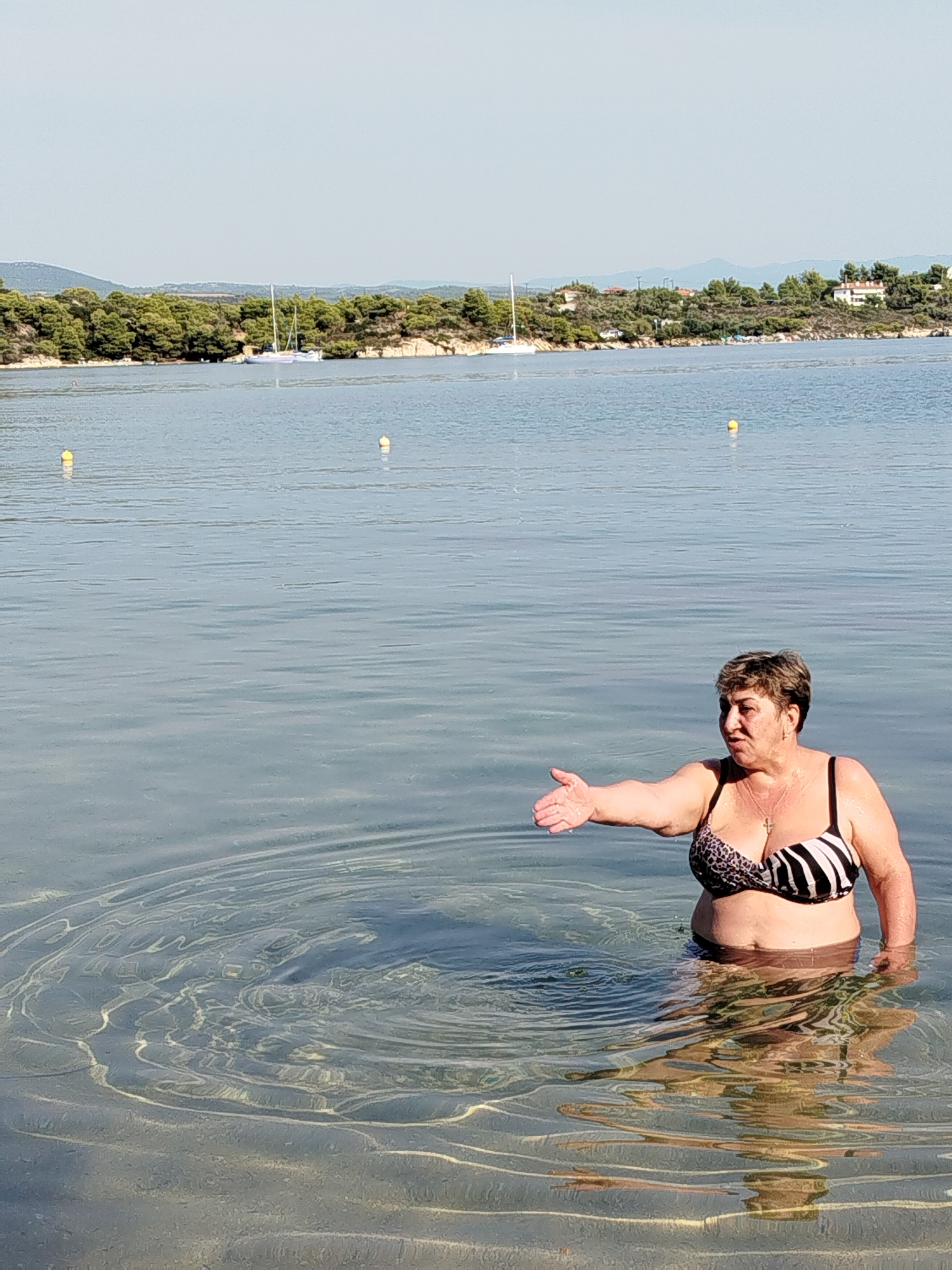 A woman wearing a black and white striped bikini with leopard print accents is standing in the clear, shallow water of a bay in Sithonia Municipal Unit, Greece. The water is calm with visible ripples and sunlight reflecting off the sandy bottom. The woman is facing the left side of the frame, extending her right arm and hand outwards, palm open, as if gesturing or reaching for something. Her gaze is directed towards her outstretched hand. Behind her, the shoreline is lined with lush green trees and scattered houses. The sky is clear and blue, with a few sailboats visible in the distance on the tranquil sea. The overall scene depicts a peaceful, sunny day at a coastal location.