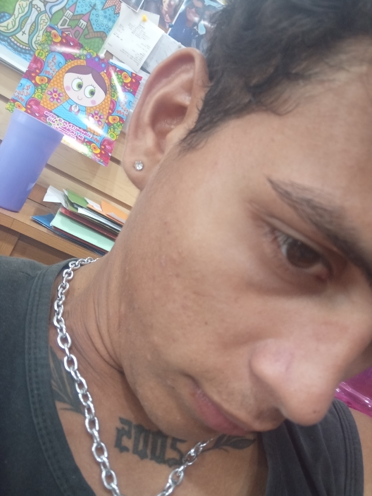 A close-up, slightly tilted selfie shows a person with light brown skin, dark hair, and brown eyes. They have a silver chain necklace and a stud earring. A "2005" tattoo is visible on their neck. The background is blurred and includes a colorful picture of a cartoon girl, a light purple cup, and papers stacked on a shelf. The location in the city of Juan Griego, Venezuela, cannot be determined from the visual information.