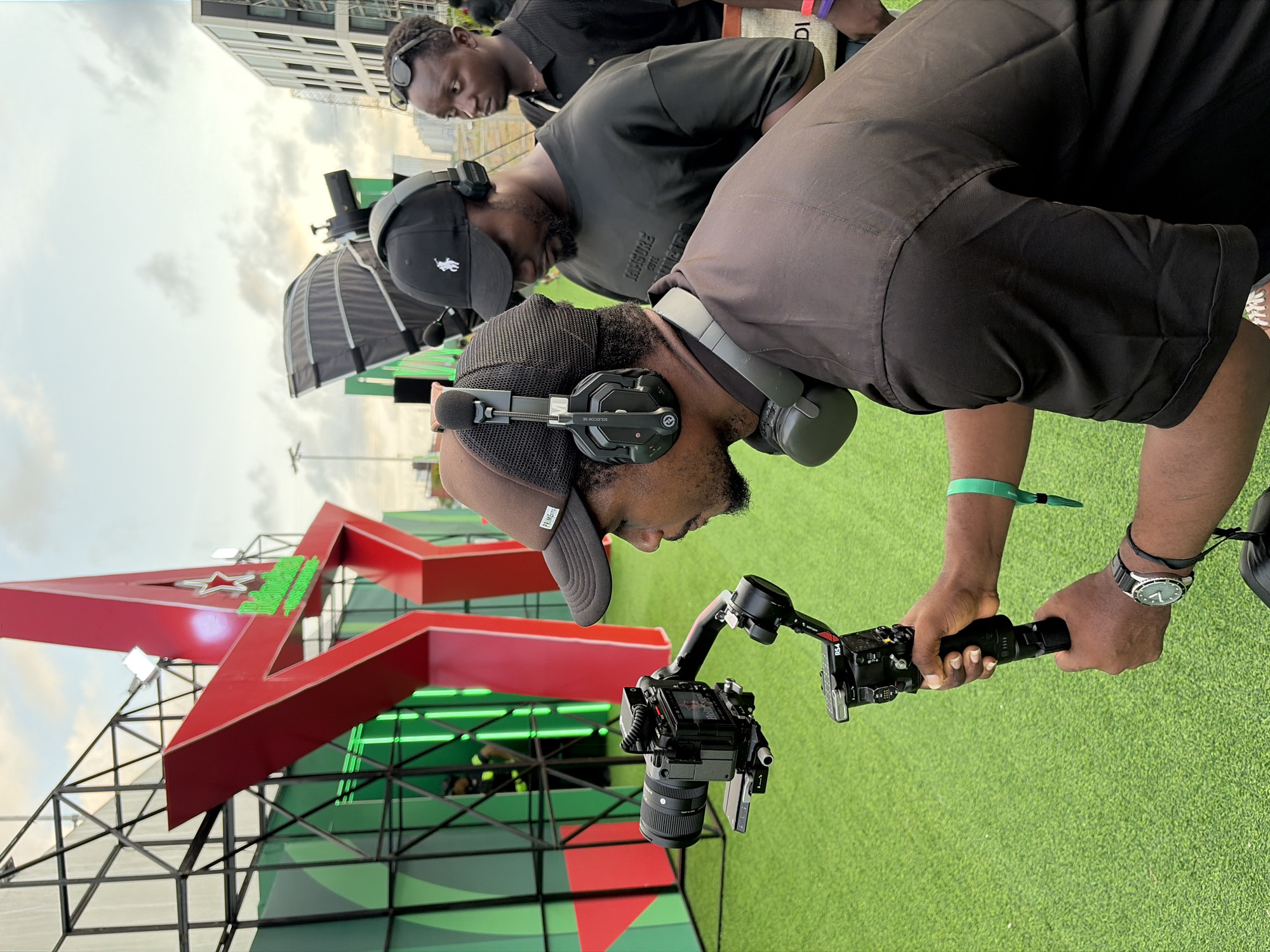 Three men are present. The man in the foreground operates a camera rig. He wears a brown cap, black shirt, watch and has headphones on. The man in the middle ground wears a polo hat and a black shirt and has headphones on. The man in the background wears sunglasses. They are in front of a red Heineken star. The setting appears to be in Lagos, Nigeria, given the Heineken sign. They stand on a green turf ground. Tall buildings are in the background.