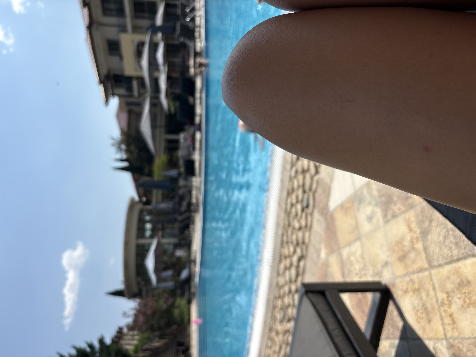The image shows a close-up view of a person's legs resting on a lounge chair, with a swimming pool and resort buildings in the background. The setting appears to be a sunny day at a resort, possibly in a warm climate. The pool is blue and appears to be outdoors. There are lounge chairs and umbrellas around the pool, and buildings that resemble hotel or resort accommodations. The focus is on the legs in the foreground, with the background slightly blurred. The overall impression is one of relaxation and leisure.
