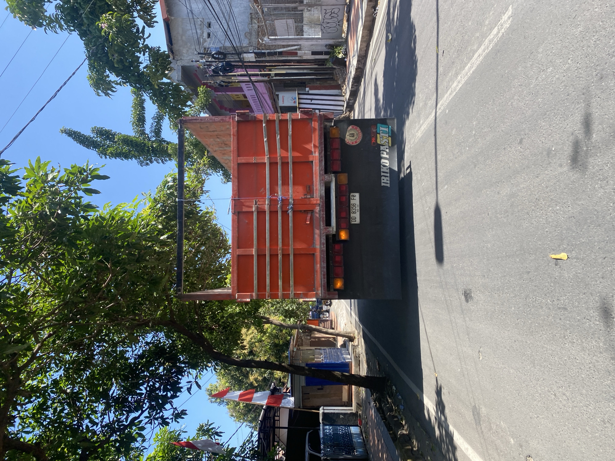 The image shows the rear of an orange truck driving on a paved road in the city of Bantaeng, Indonesia. The truck has the license plate "DD 8356 FO". The road is lined with trees and buildings. There are two Indonesian flags hanging from poles on the left side of the road. The sky is clear and blue. The overall scene depicts a typical street view in an Indonesian city.