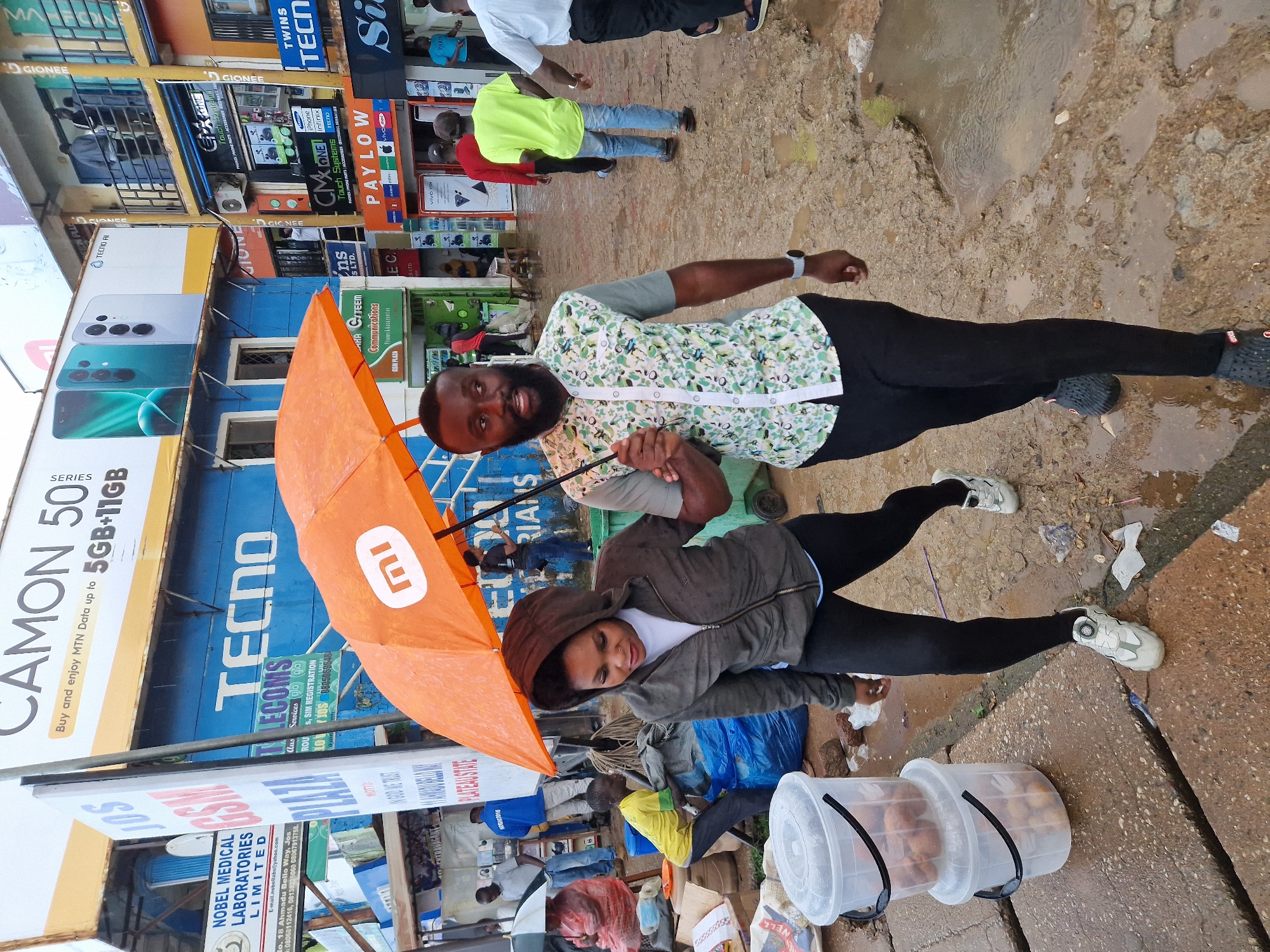 A medium shot captures a couple walking under an orange umbrella in an outdoor market setting. The man, in the foreground and to the right, wears a patterned short-sleeved shirt and dark pants, holding the umbrella. Beside him, a woman in a hooded jacket and black leggings walks to his left. Both are looking forward, suggesting they are walking together. The ground is wet and muddy, indicating recent rain, with puddles visible. In the background, various shops with signs like "TECNO," "GSMPLAZA," and "CMX One" are visible, suggesting a commercial area. Several other people are present in the background, some walking and others standing. Two clear plastic buckets filled with small, round items, possibly fruit or vegetables, are placed on the cracked concrete ground in the lower left of the frame. The lighting suggests it is daytime, possibly overcast due to the presence of the umbrella.