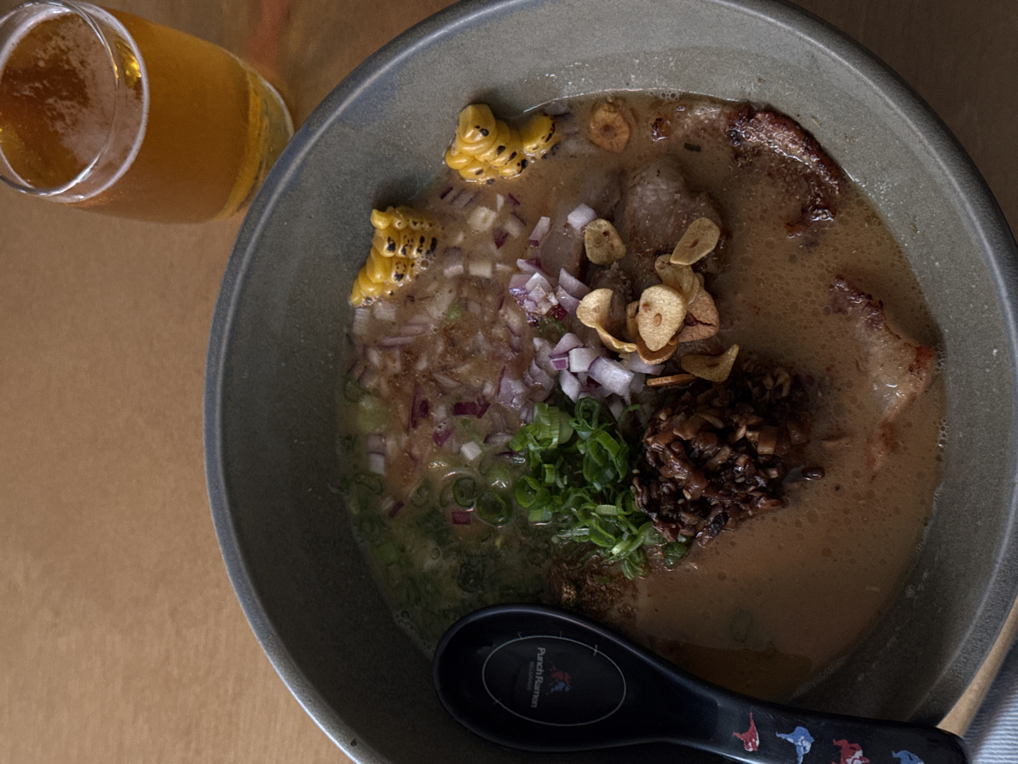 A high-angle, indoor shot presents a bowl of ramen alongside a glass of beer on a tan-colored table surface. The ramen bowl, made of gray ceramic, is filled with broth and toppings: grilled corn on the cob, diced red onions, sliced green onions, fried garlic chips, and a scoop of dark brown mushrooms. A black spoon rests in the bowl. The beer glass is partially visible in the top left corner, filled with a light-colored brew. The location is Punch Ramen in Belgrade, Serbia, as evidenced by the restaurant's logo printed on the black spoon.