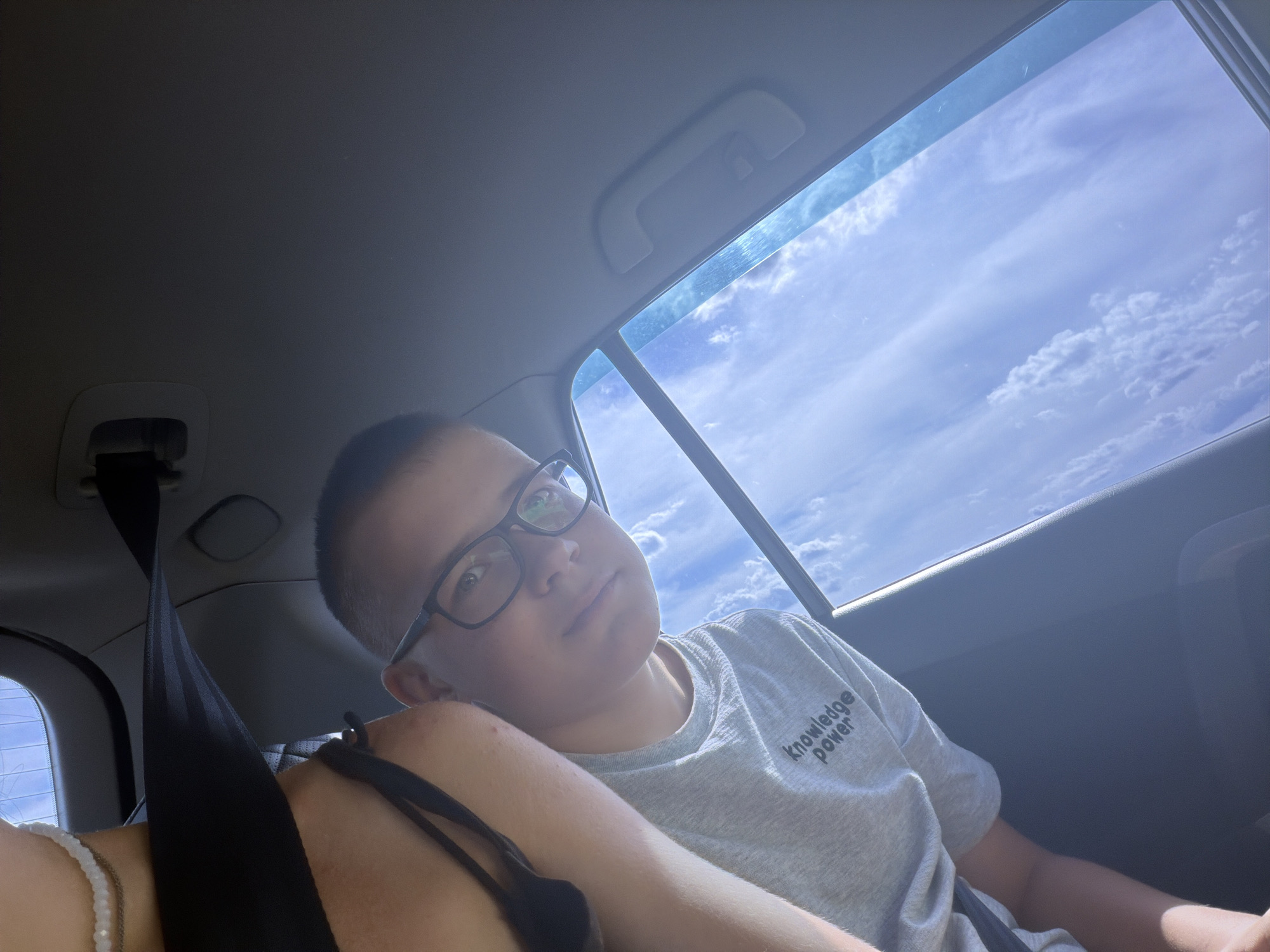A young boy wearing glasses and a grey t-shirt with the words "knowledge power" is sitting in the back seat of a car. The boy is looking up and to the side, with his left arm resting on someone's shoulder. The car's interior is visible, including a seatbelt and the roof lining. Through the car window, a blue sky with scattered clouds can be seen. The scene appears to be a snapshot taken from inside the vehicle, possibly during a drive through the city of Novoe Teneevo, Russia.