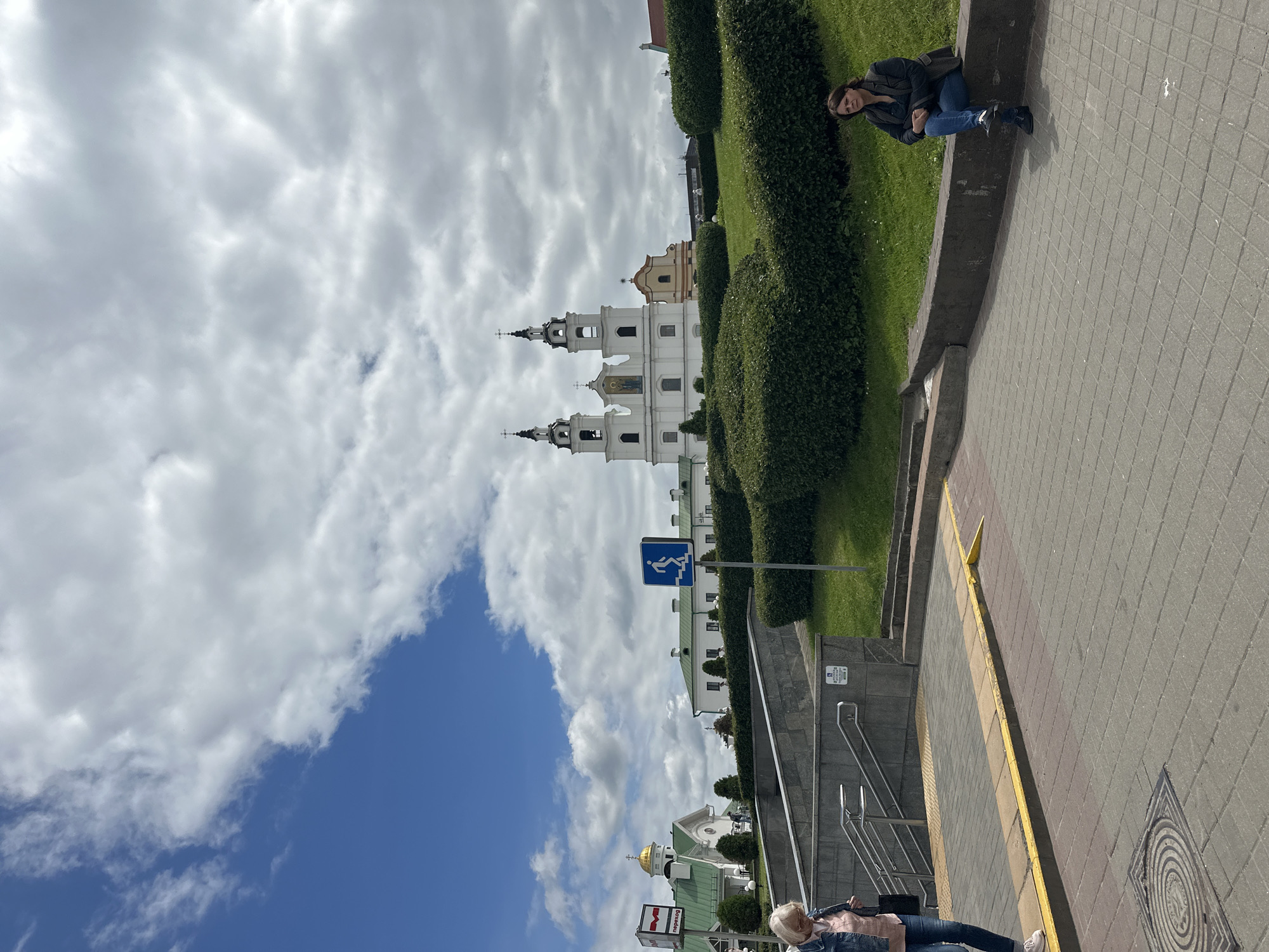 The image depicts a street scene in Minsk, Belarus, with a large white church featuring two prominent towers and crosses as the central architectural element. A smaller building with a golden dome is visible to the left of the church. The foreground shows a paved walkway with a yellow tactile warning strip, leading to a set of stairs descending underground. Two women are present in the scene: one, wearing a denim jacket and pink top, is walking down the stairs, and the other, dressed in a dark jacket and jeans, is seated on a low concrete wall on the right side of the frame. The background is dominated by a bright blue sky with scattered white clouds and lush green bushes that form manicured hedges. A blue sign with a white icon of a person descending stairs is placed near the hedges. The overall environment suggests a public space, possibly near a historical or religious site.