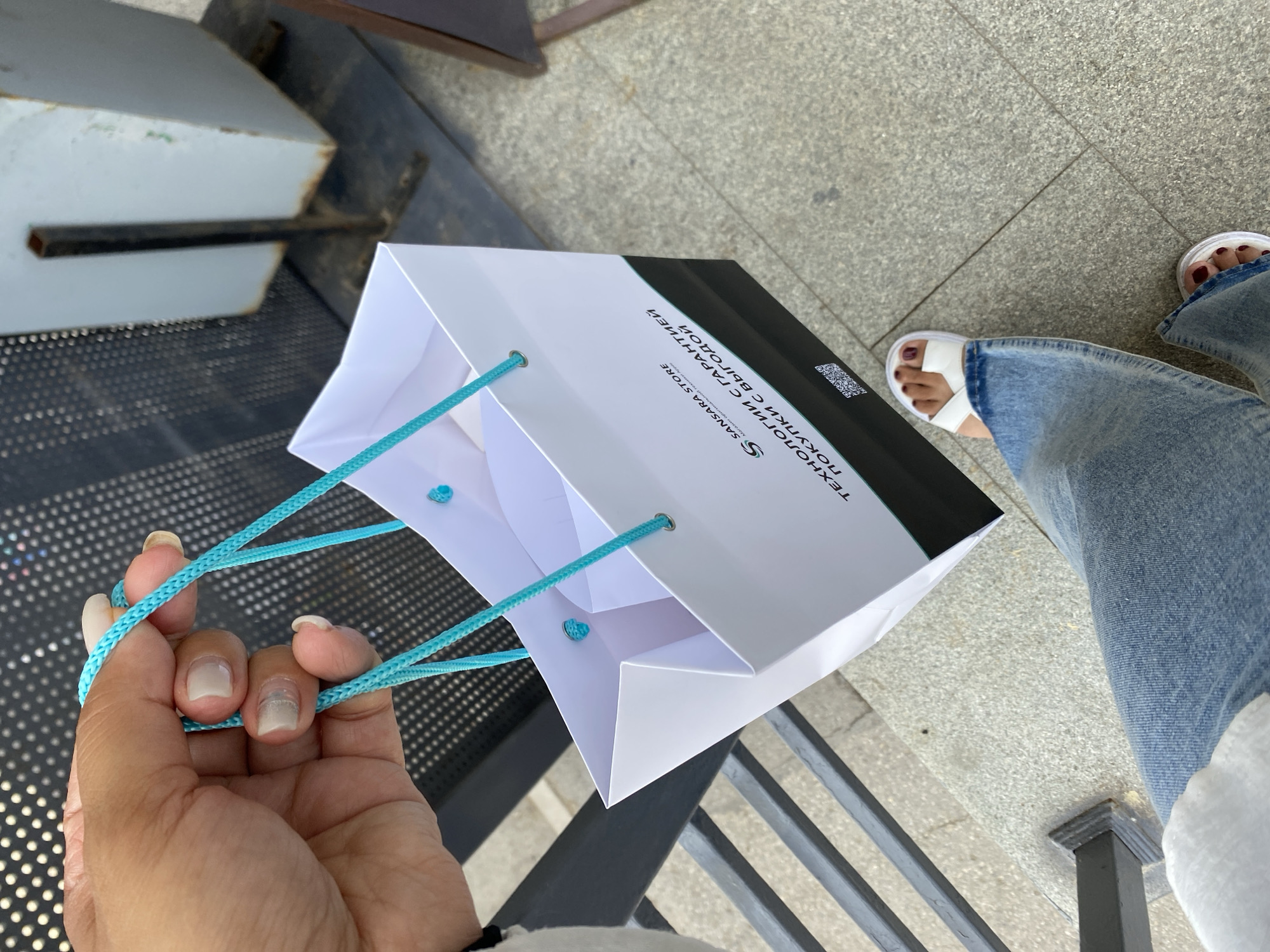 A person's hand with manicured nails holds the turquoise rope handles of a white paper shopping bag. The bag has black lettering on its side, which includes "SANSARA STORE" and a QR code. The person is wearing blue jeans and white sandals, visible at the bottom of the image. The background suggests an outdoor urban environment with a metal railing and textured pavement. The specific location in Ufa, Russia, cannot be determined from the visual information provided.